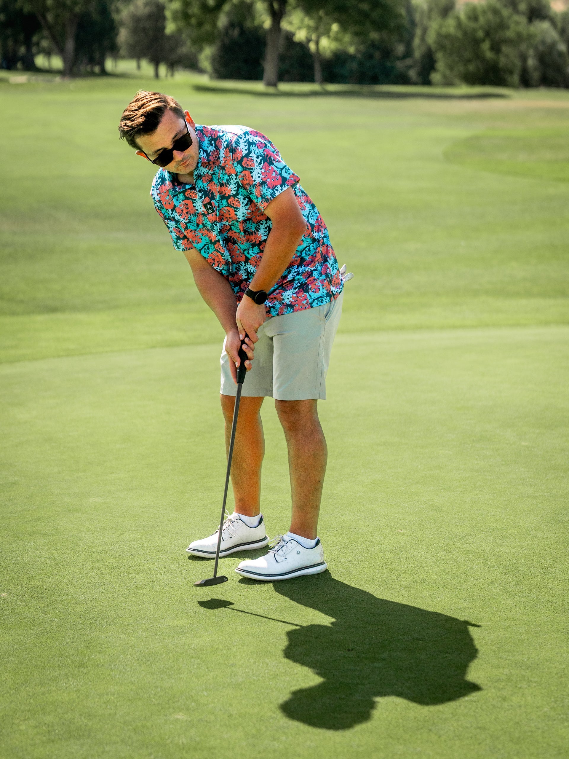 Man in colorful tropical golf shirt, beige shorts, white golf shoes, sunglasses, putting on a green golf course.