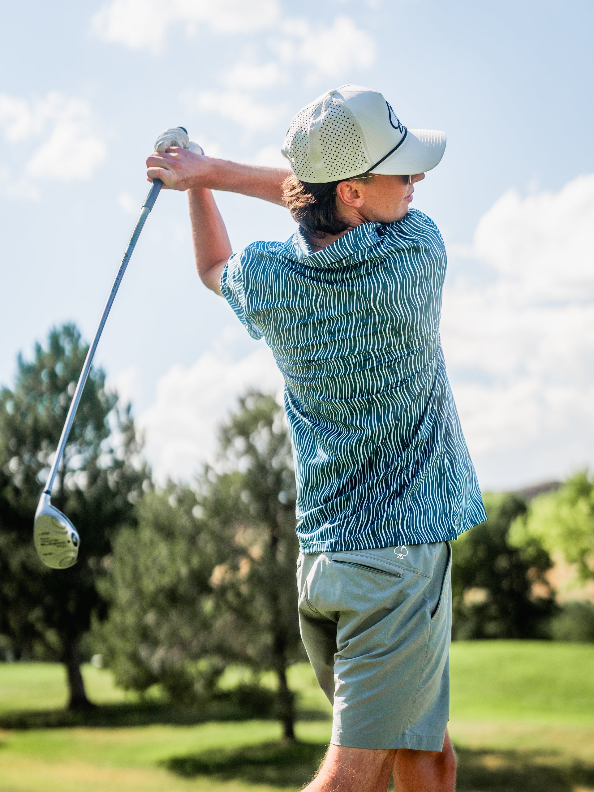 Male golfer in a blue patterned shirt, beige shorts, white cap, swinging golf club on a sunny course.