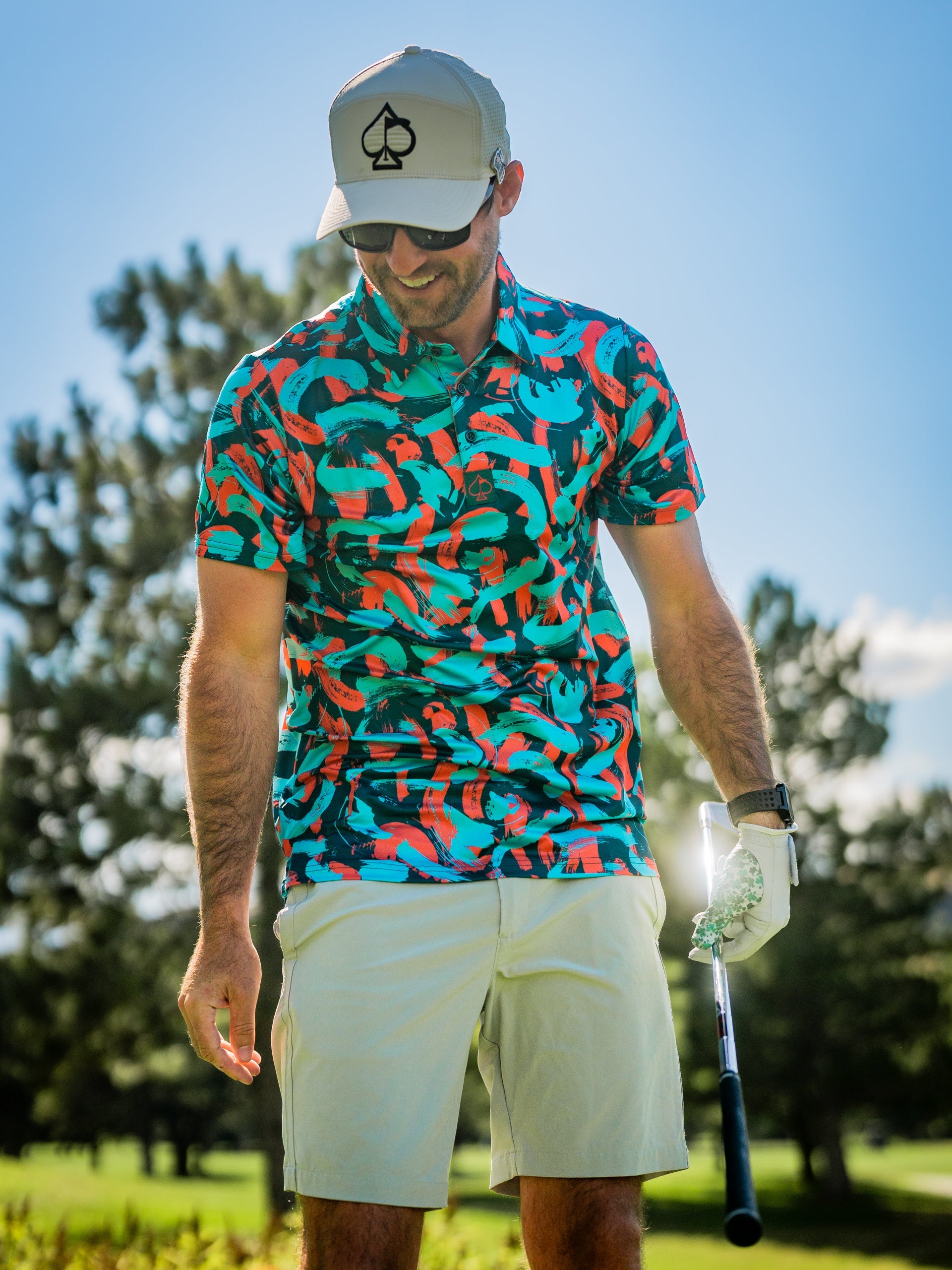 Man wearing a vibrant Paint Strokes Coral golf shirt on the course, holding a golf club with a sunny backdrop.