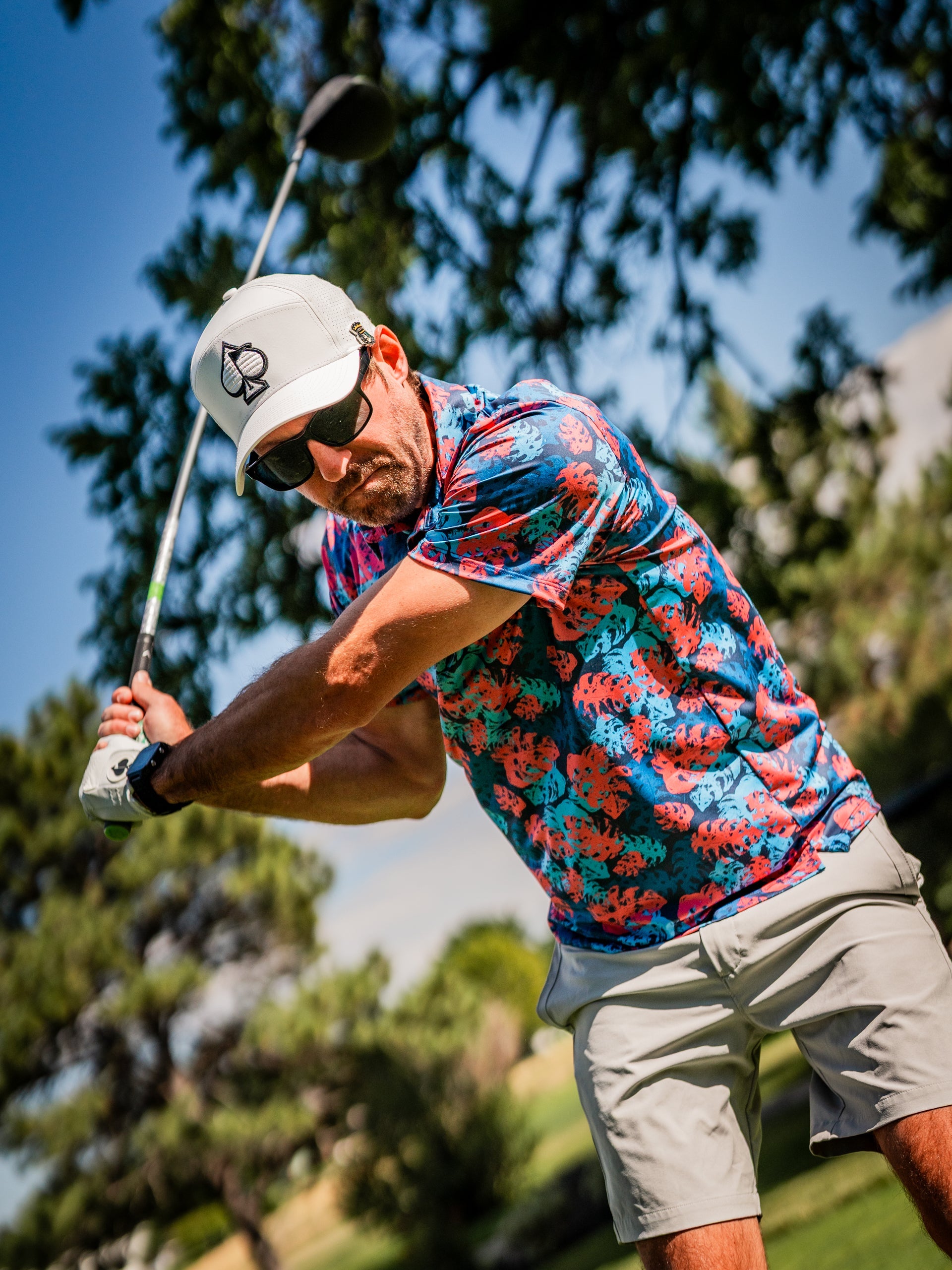 Male golfer in colorful tropical shirt, white cap with logo, dark sunglasses, swinging golf club on sunny course.