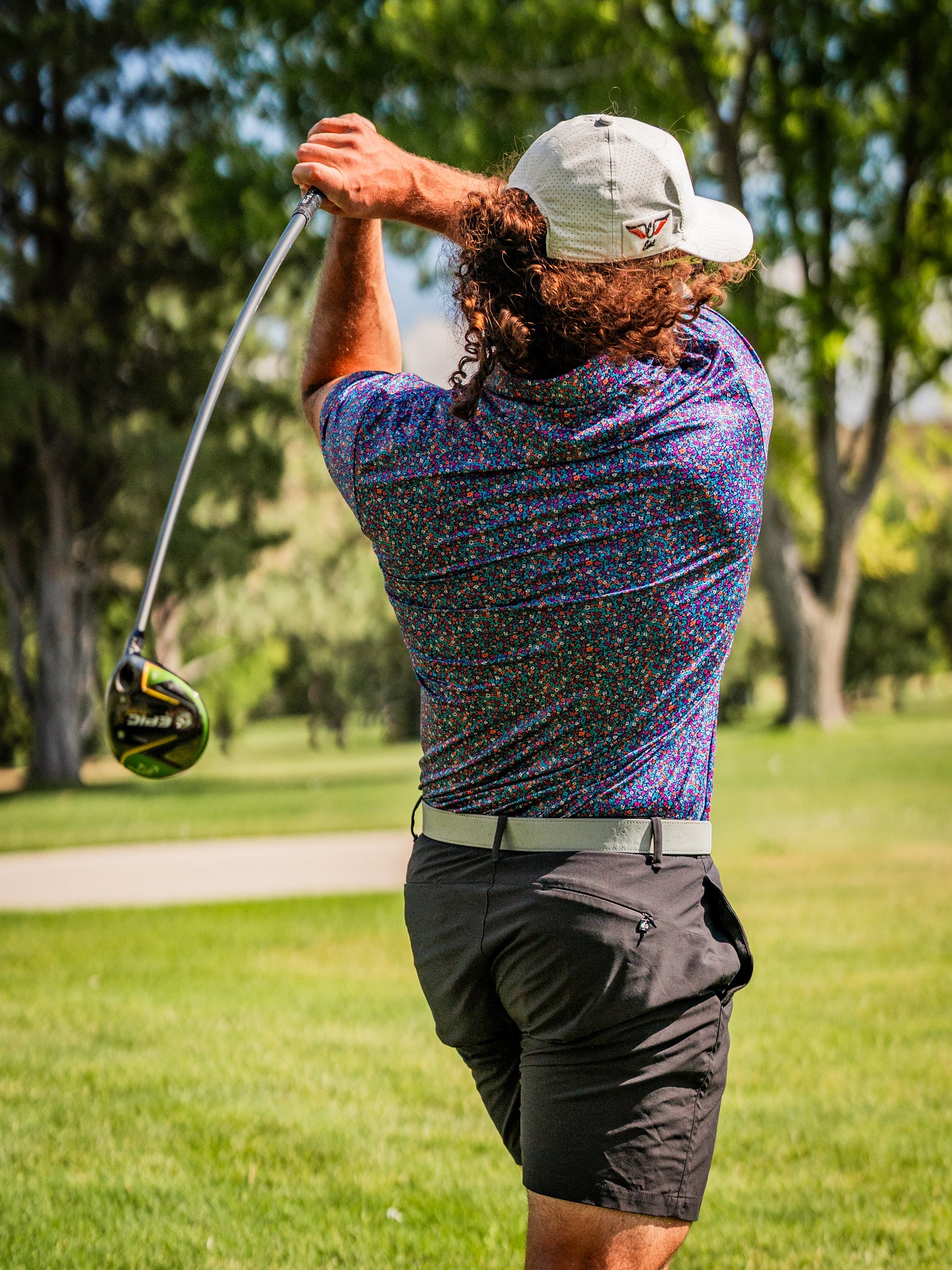 A golfer swings a driver while wearing a vibrant lavender floral-patterned shirt and black shorts on a sunny course.
