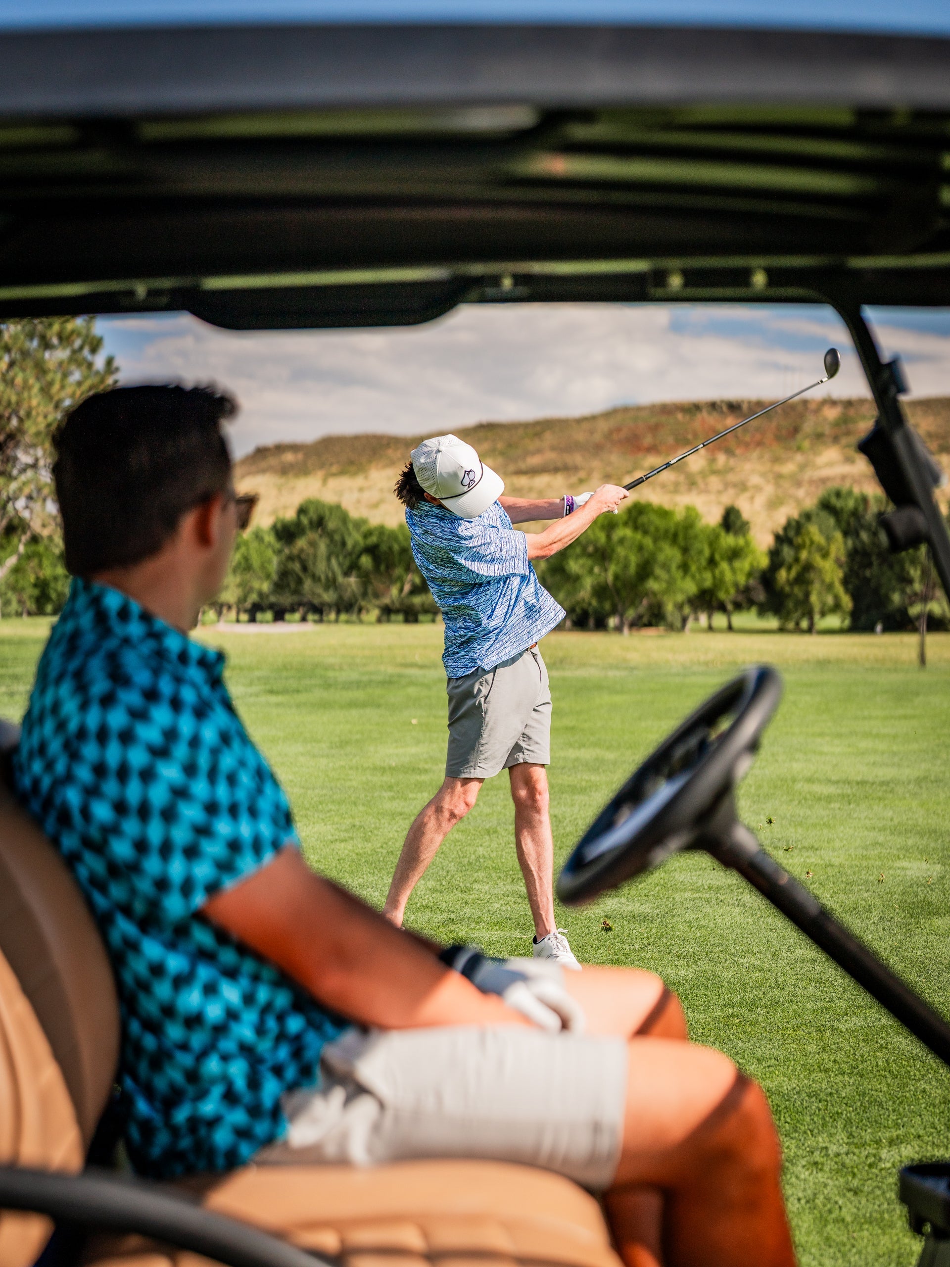 A golfer in a wavy checkered shirt swings a club while another golfer relaxes in a golf cart on the course.