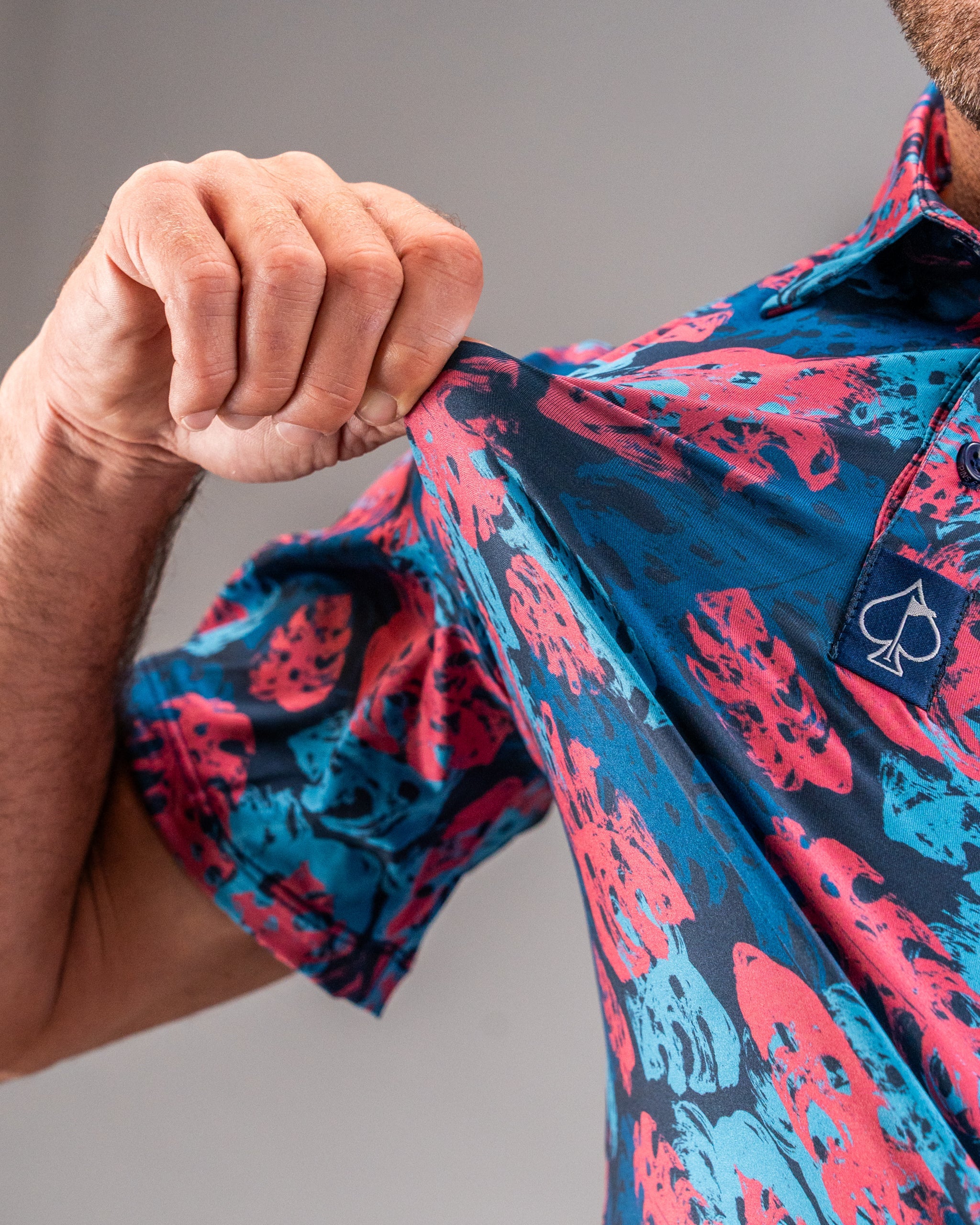 Close-up of a man wearing a vibrant, patterned golf shirt in blue, pink, and red with a spade logo, showcasing bold golf a...