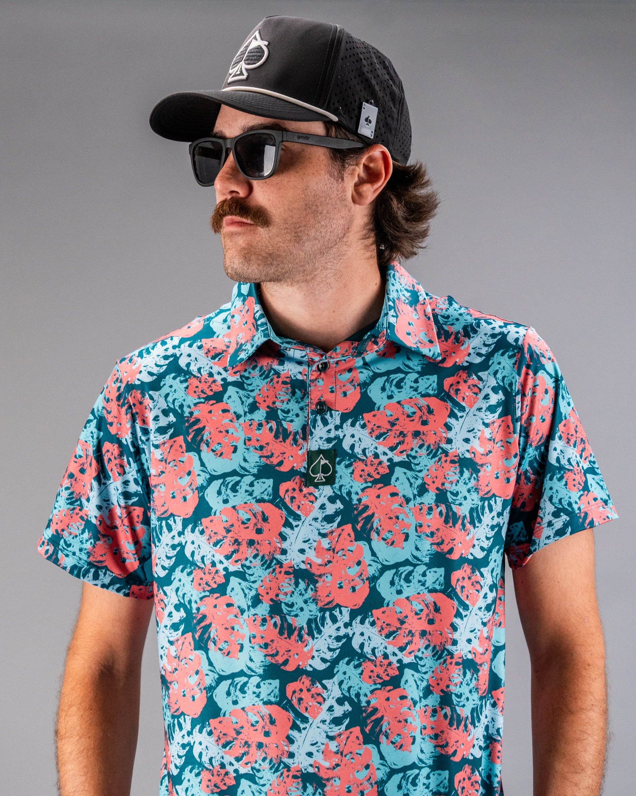Man wearing a tropical patterned shirt with pink, blue, and teal leaves, paired with a black cap and sunglasses.