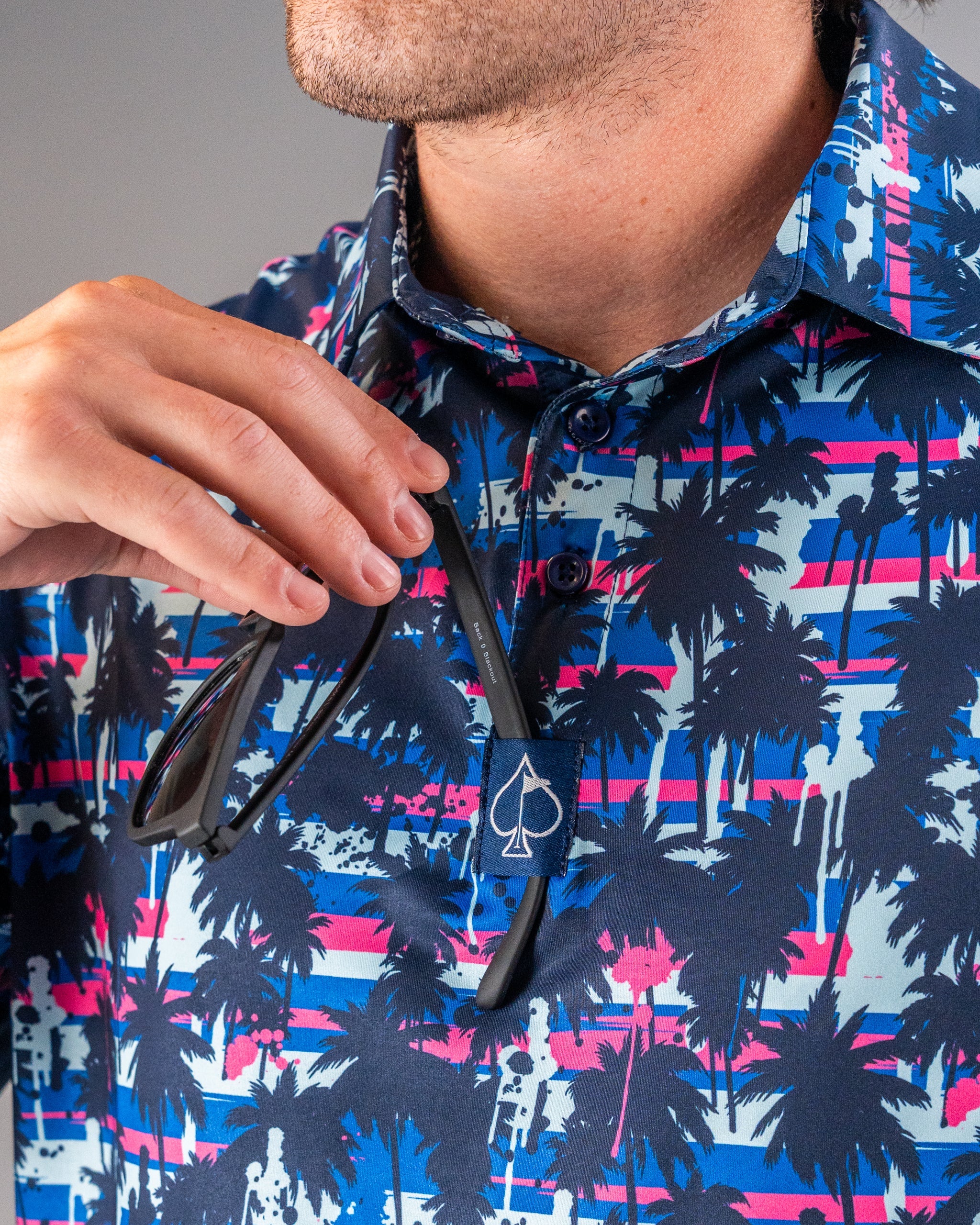 Close-up of a man in vibrant tropical golf shirt holding stylish black sunglasses with bold palm tree print, perfect for s...