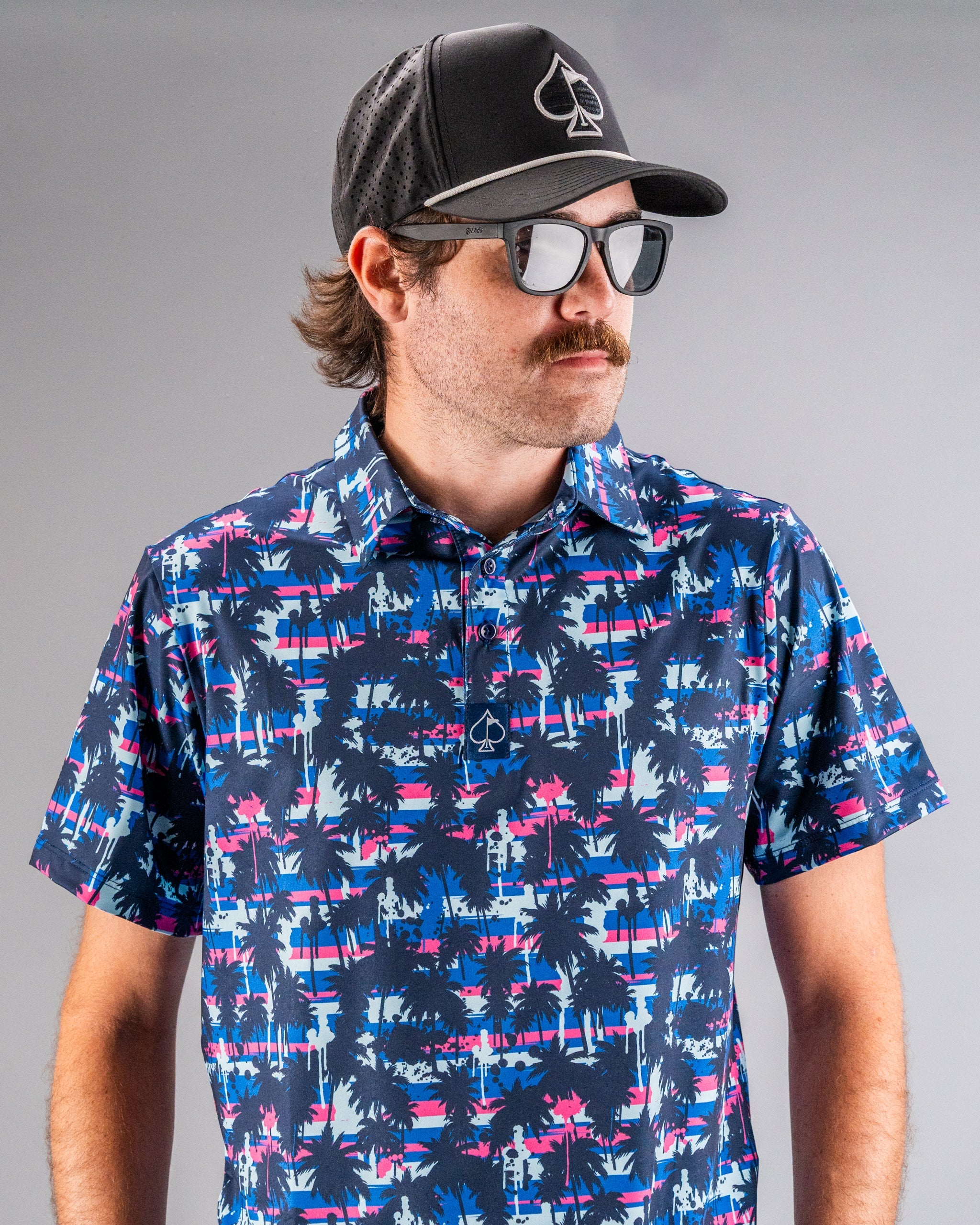 Man wearing a black cap with a spade logo, dark sunglasses, and a colorful tropical print golf shirt featuring navy, pink,...