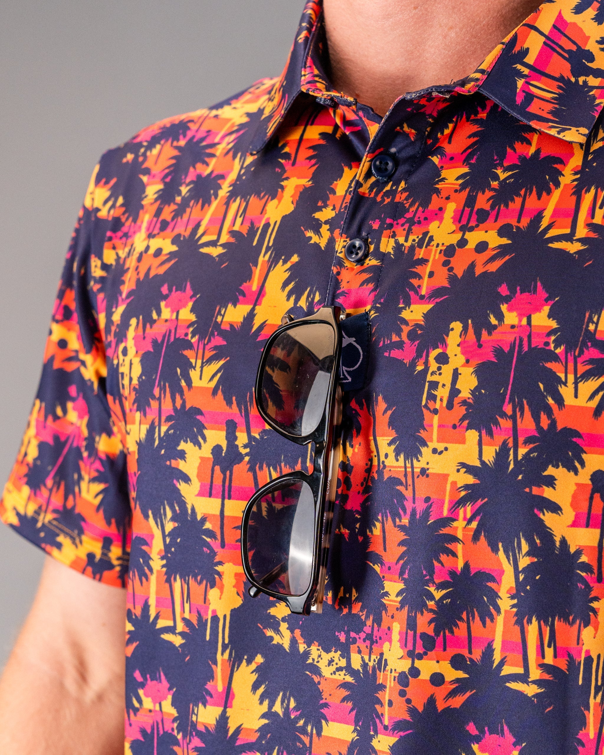 Bright tropical palm tree pattern in orange, pink, and navy on a casual collared shirt with sunglasses hanging from the fr...