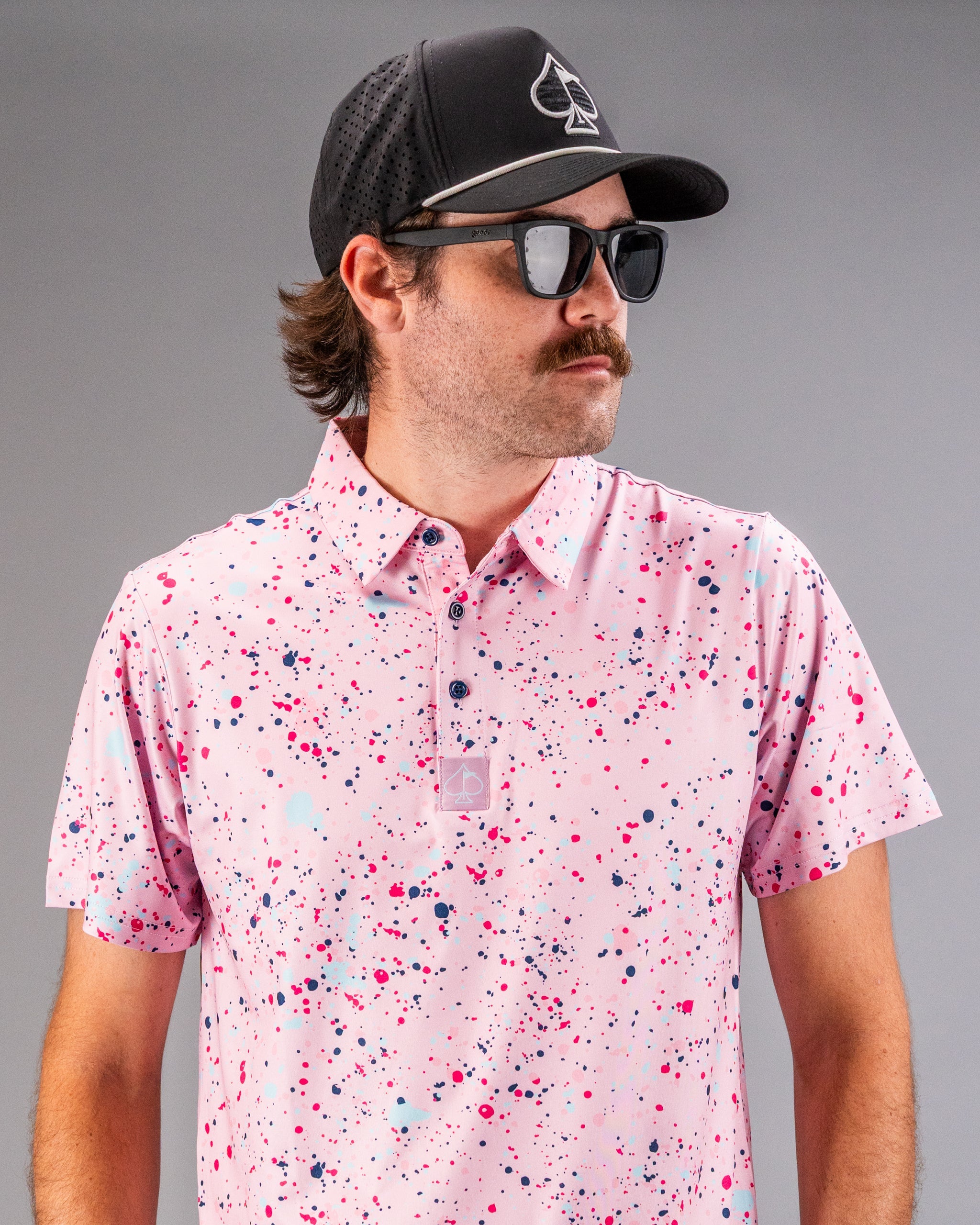 Stylish golfer wearing a pink splatter polo shirt with a black cap and sunglasses, showcasing bold golf fashion.