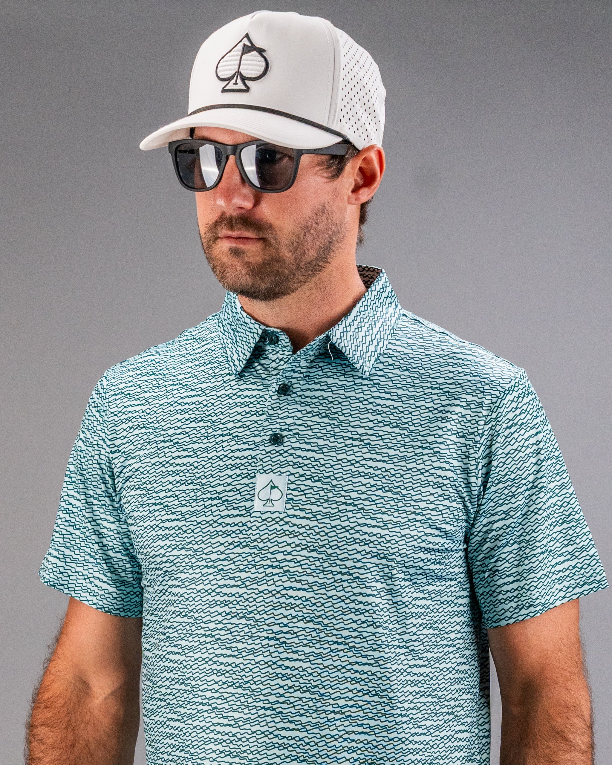 Man wearing a teal golf polo with a squiggle pattern and a white cap, showcasing a stylish and bold golf look.