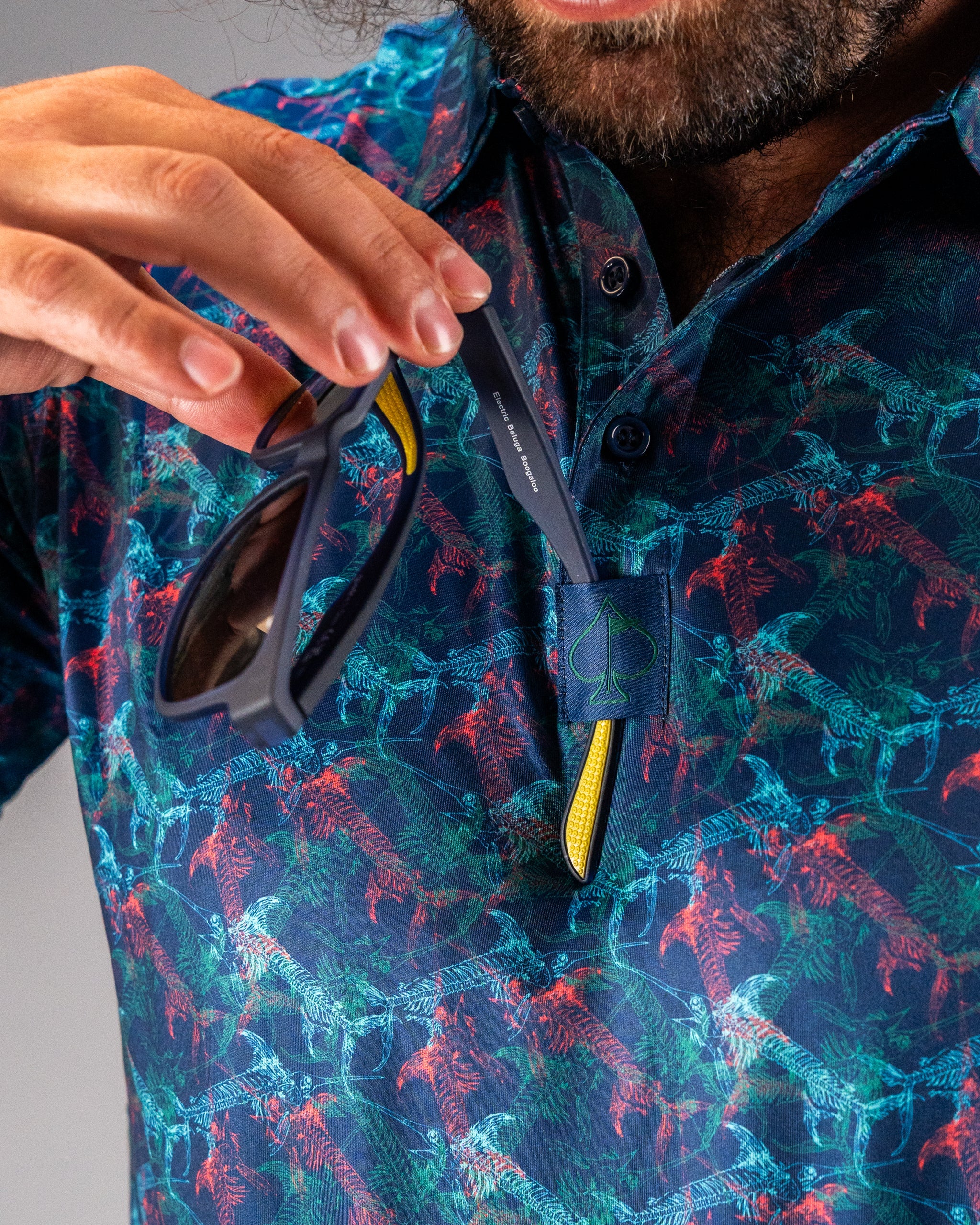 A man holds sunglasses while wearing a vibrant, patterned golf shirt featuring a swordfish design.