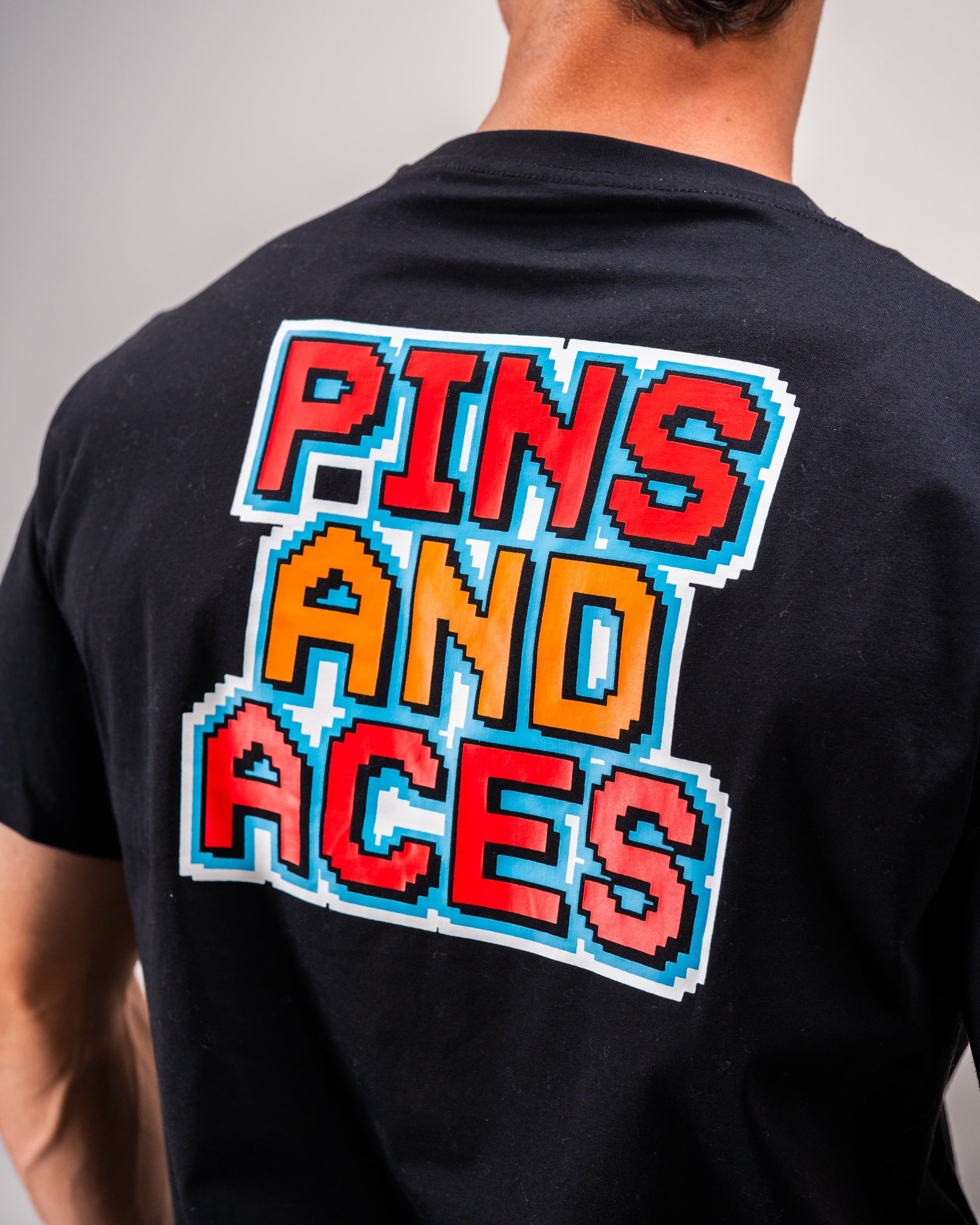 Rear view of a person wearing a black t-shirt with a colorful "PINS AND ACES" text in 8-bit font style printed on the back.