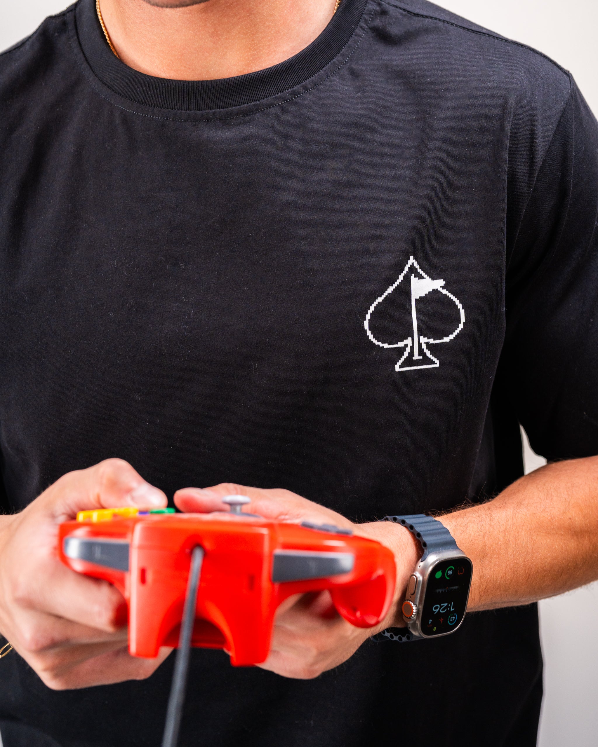 Man wearing a black t-shirt with a white 8-bit spade symbol, holding a red video game controller, and wearing a smartwatch.
