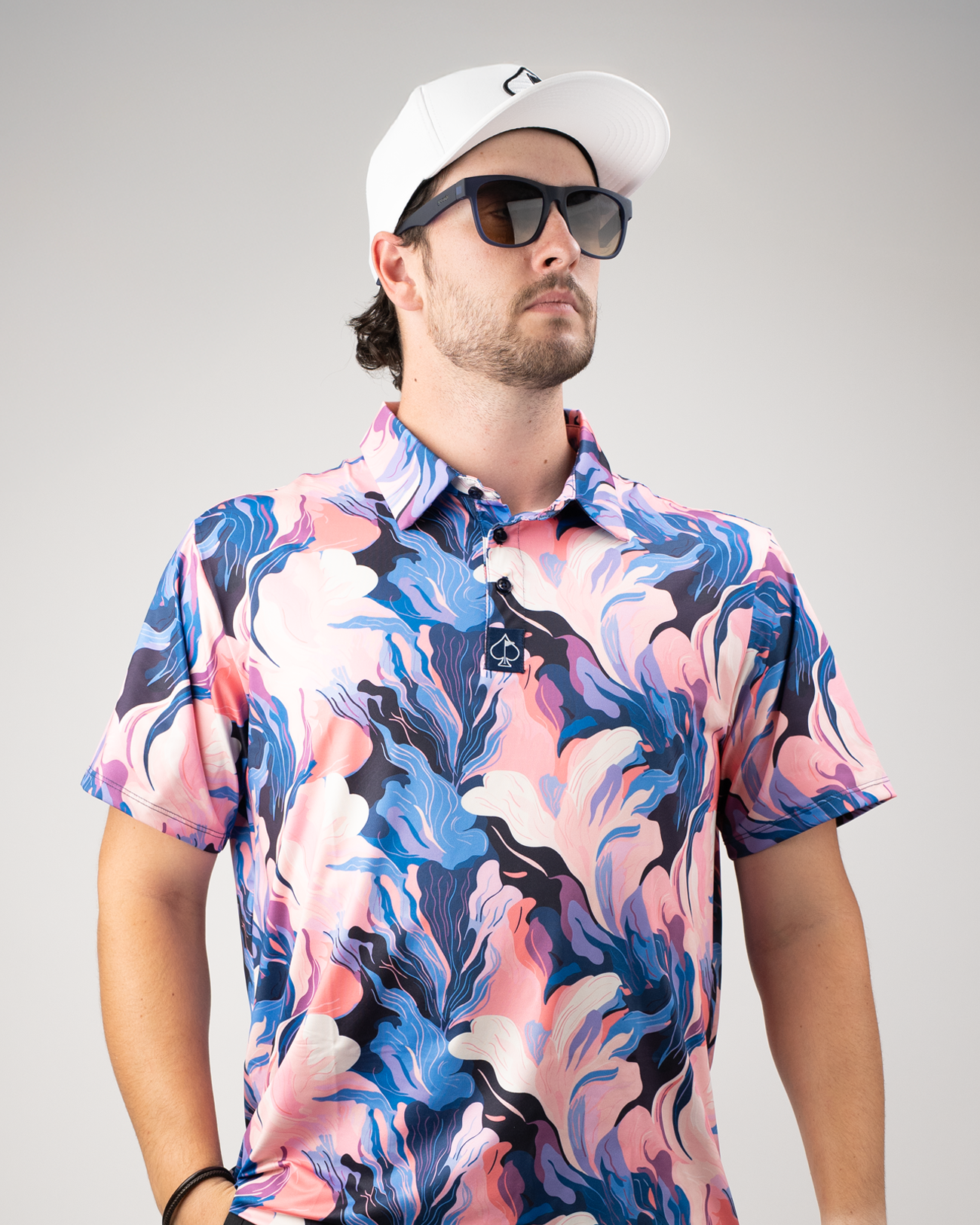 A man models a vibrant Melting Floral golf polo featuring bold pink, blue, and black abstract patterns, paired with sunglasses and a white cap.