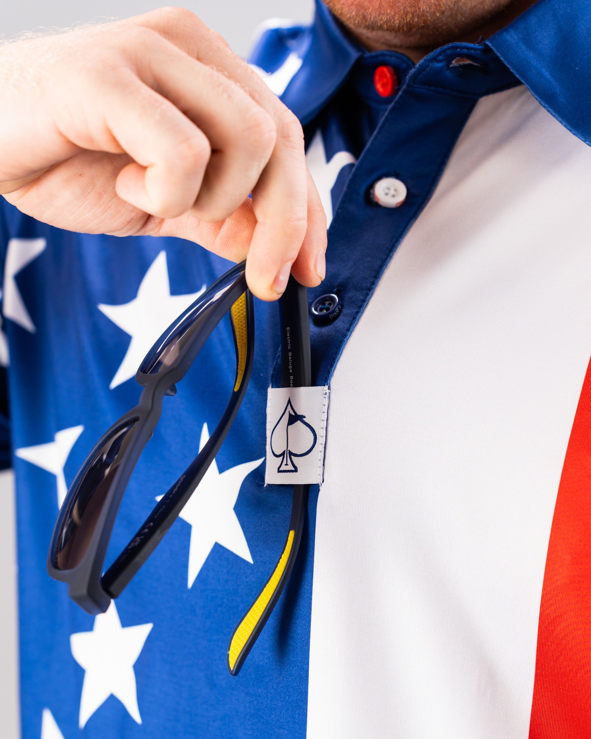 Close-up of a person holding sunglasses near a white tag with a black spade symbol, on a patriotic shirt with stars and stripes.