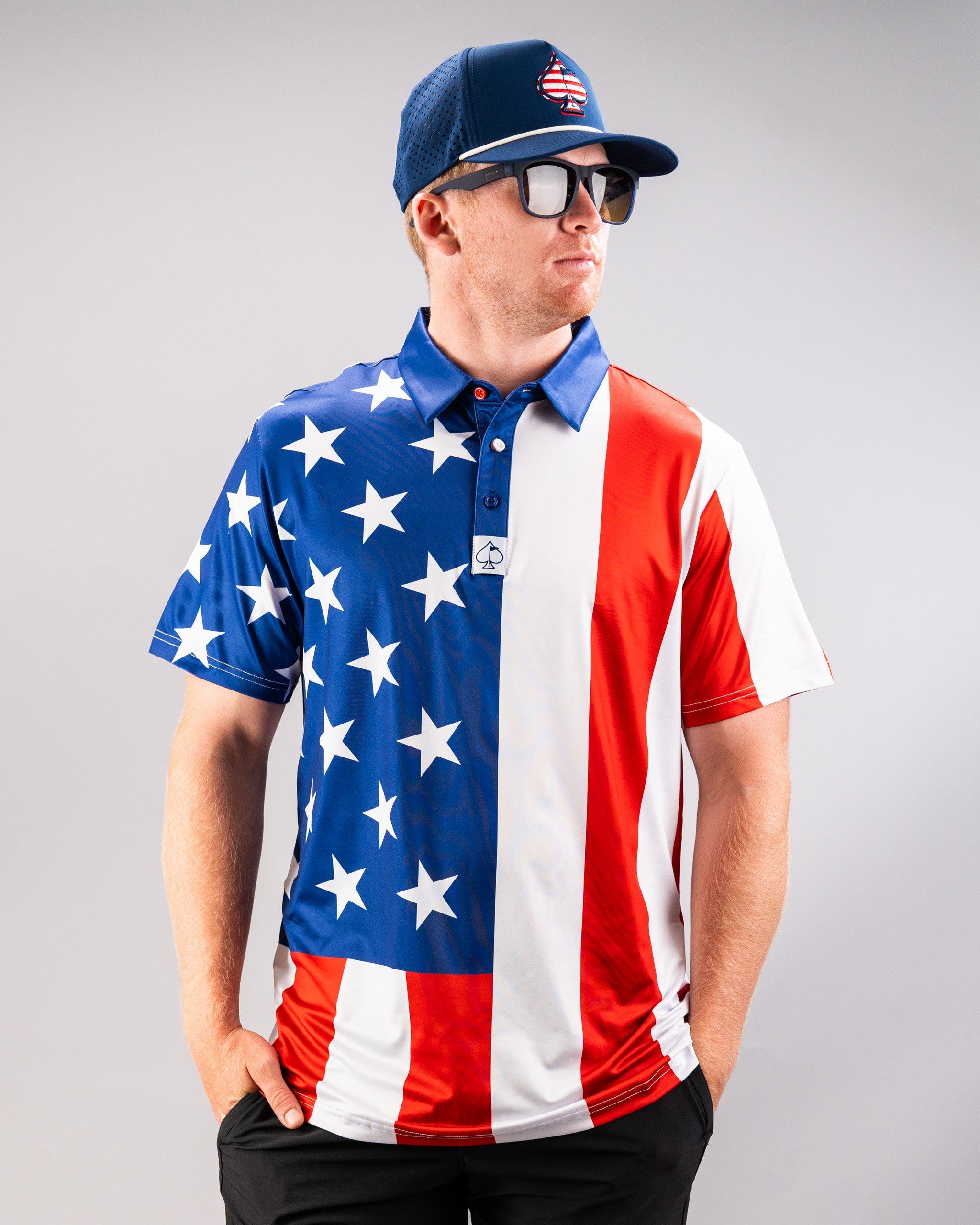 Man wearing a patriotic golf shirt with an American flag design, a blue cap with a spade logo, and sunglasses, standing against a plain background.