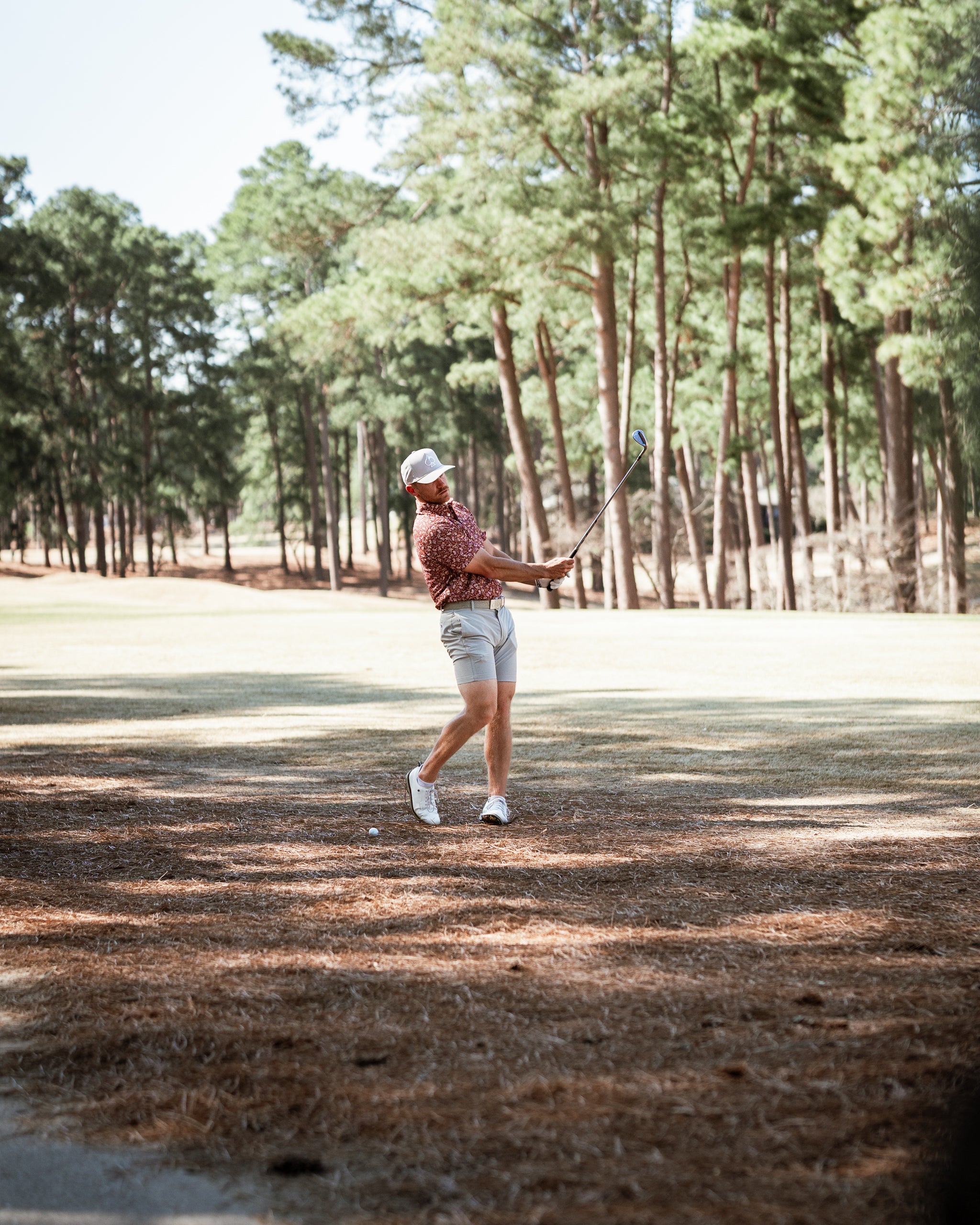A golfer swings a club on a sunlit course surrounded by tall trees, wearing the Autumn Bloom golf shirt.