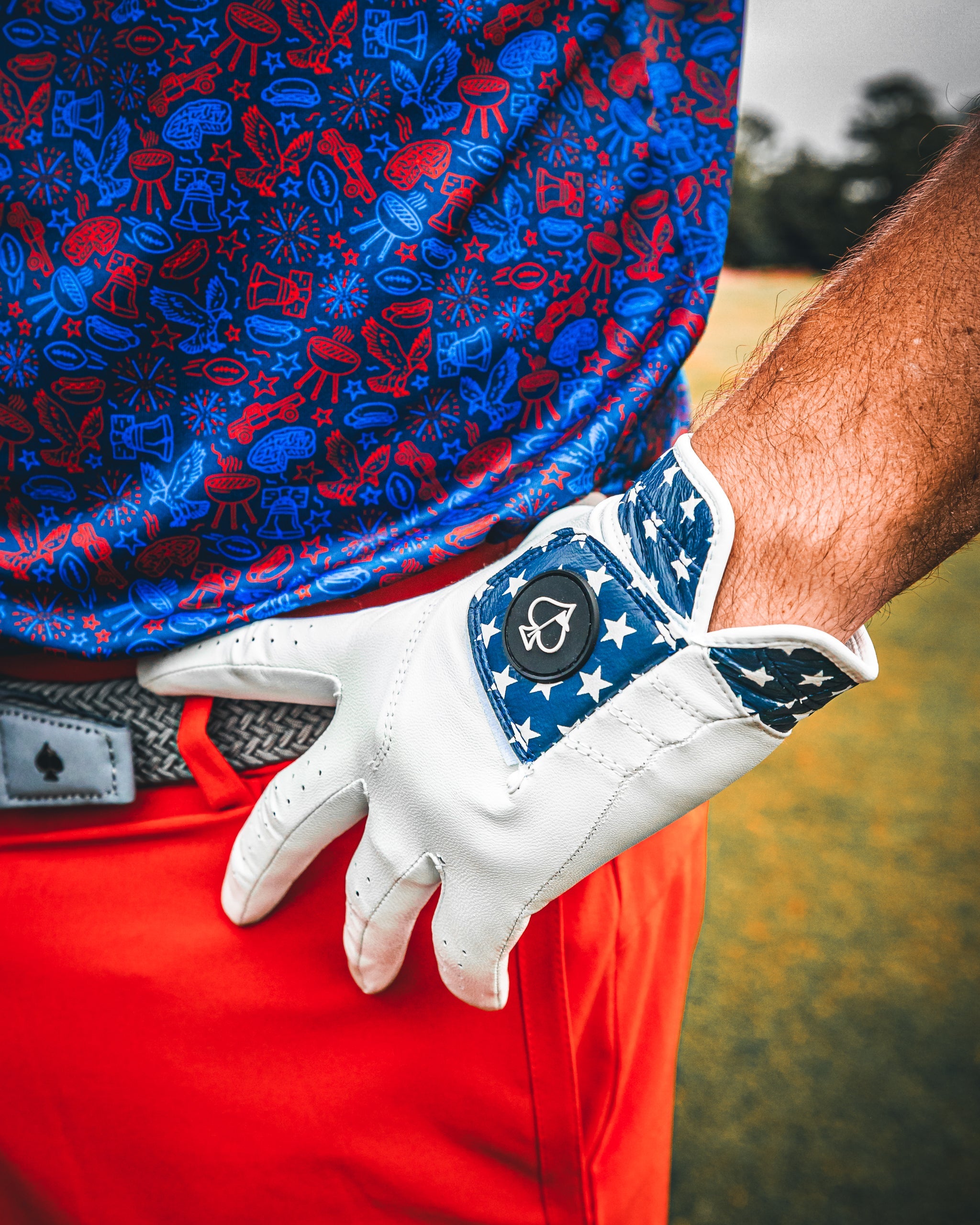 A golfer wearing a navy and red patterned shirt and bright orange pants adjusts a white golf glove with a star design.