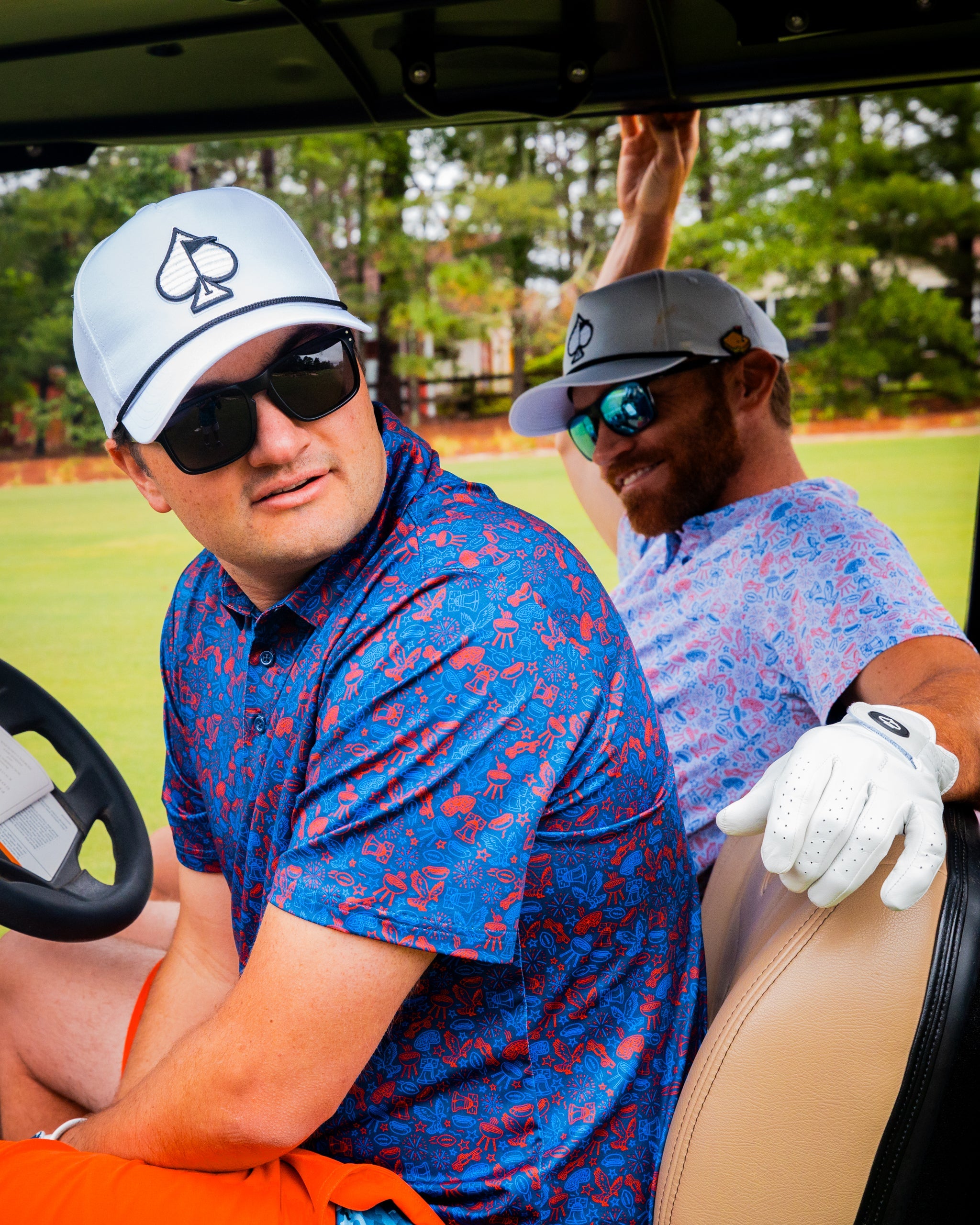 Two golfers in a golf cart, one wearing a Freedom Doodle Navy shirt and a white cap, the other in a patterned shirt and sunglasses.