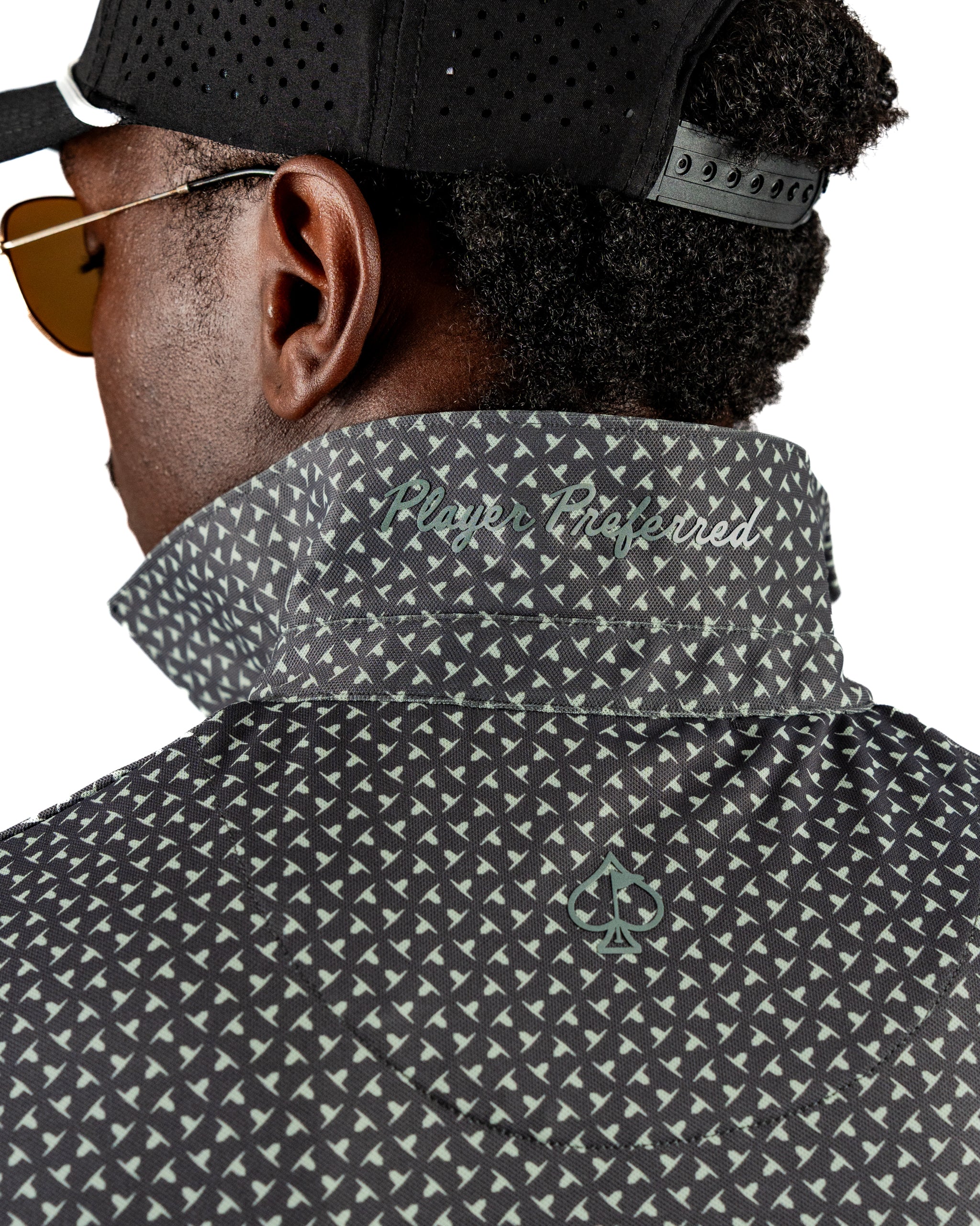 Close-up of a golfer wearing a black patterned jacket with a high collar, featuring the text "Player Crafted" and a spade logo.