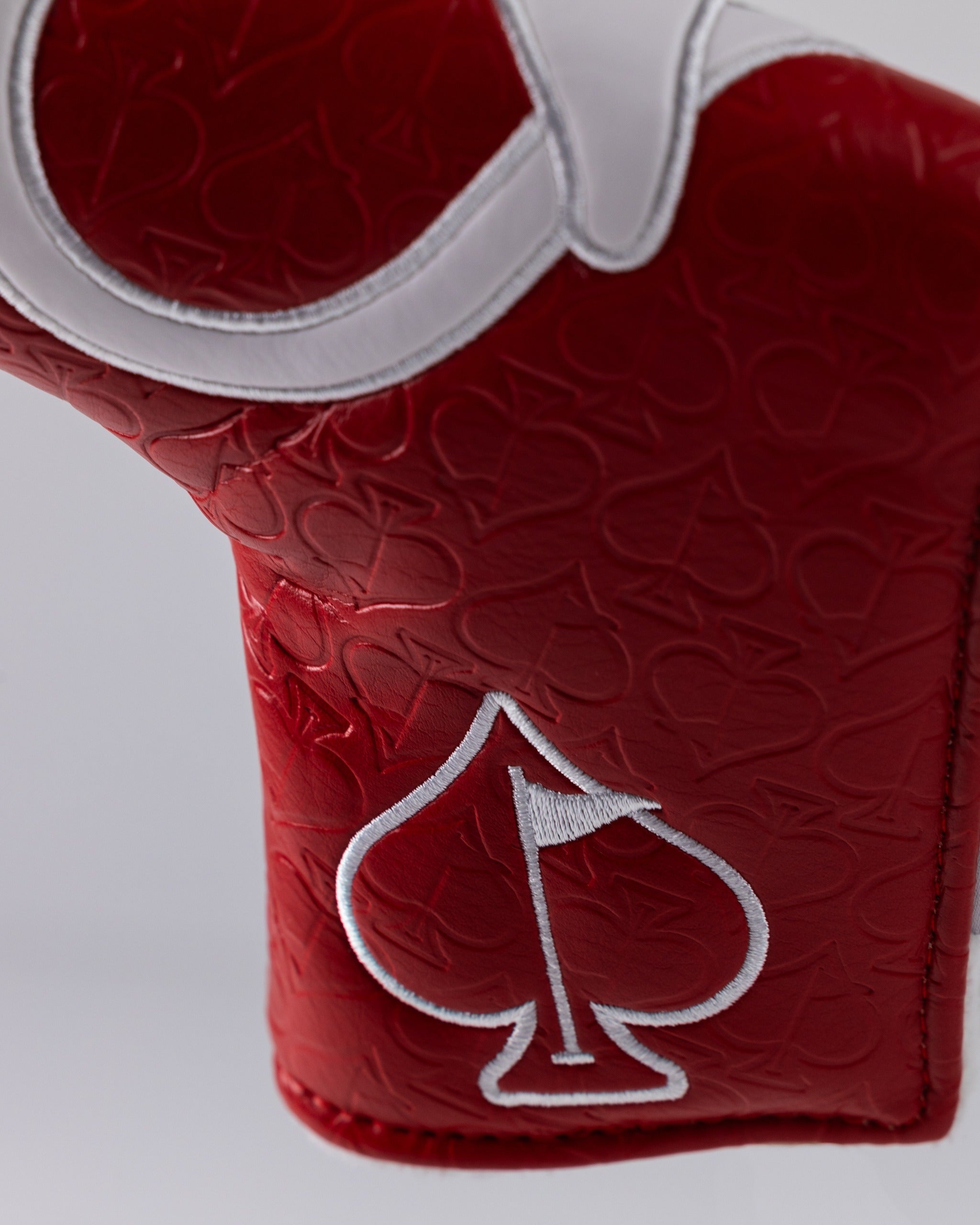 Close-up of a red golf headcover with white embroidered spade logo, textured pattern, and white trim, for bold, unique gol...
