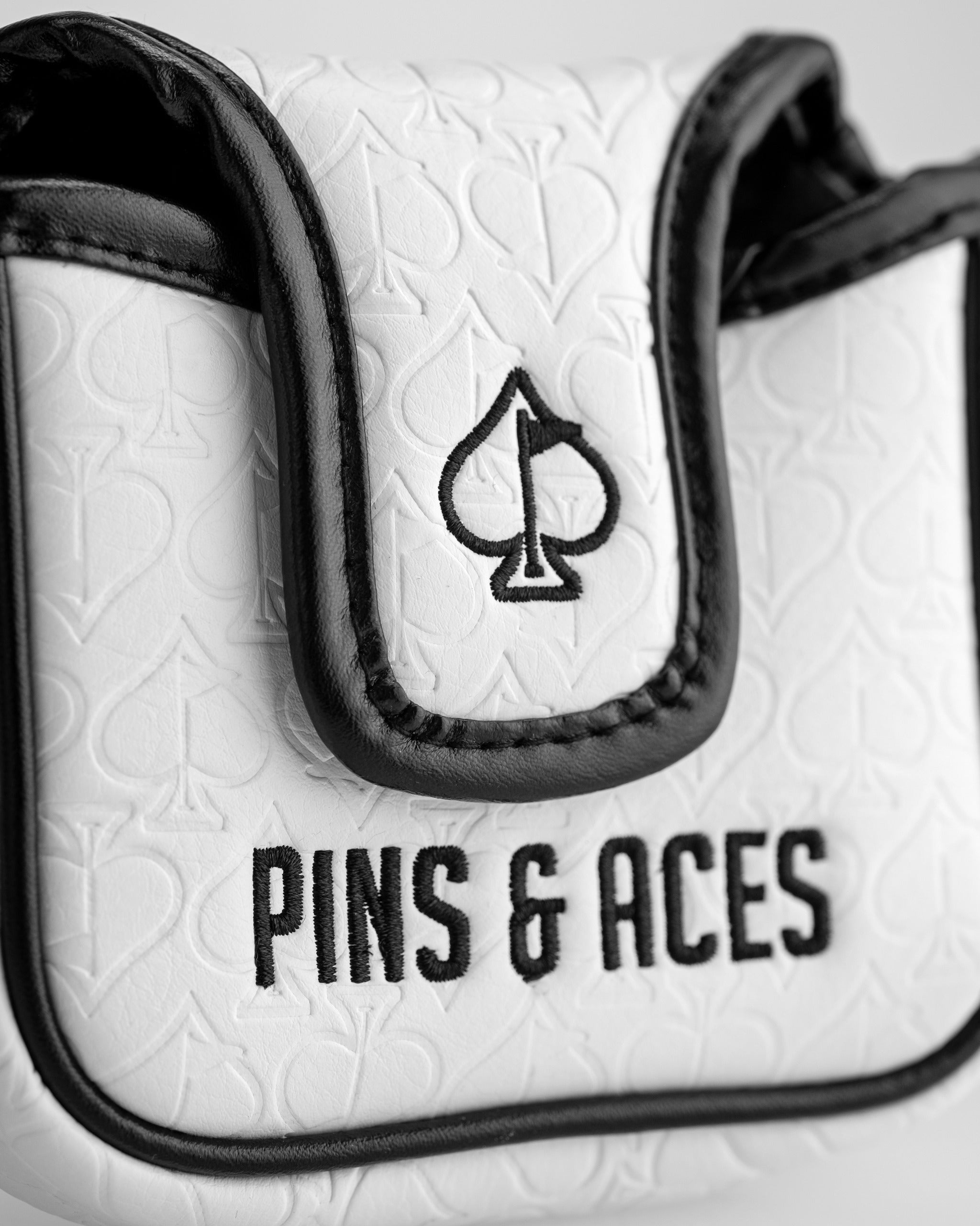 Close-up of a white golf headcover with black leather accents, embroidered spade logo, and "PINS & ACES" text, featuring a...