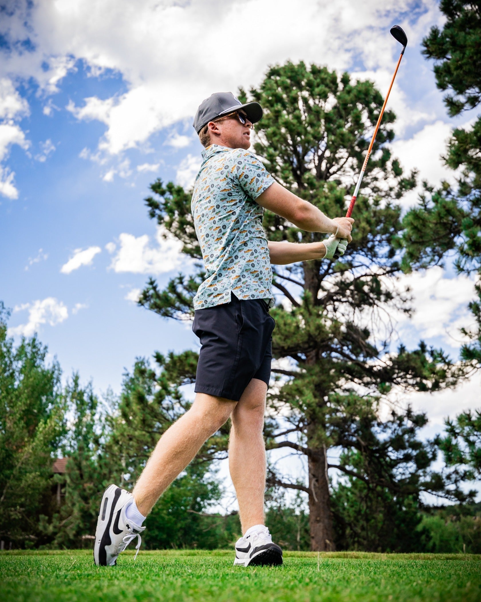 A golfer swings a club on the course, wearing a colorful patterned shirt and shorts, with trees and blue sky in the background.