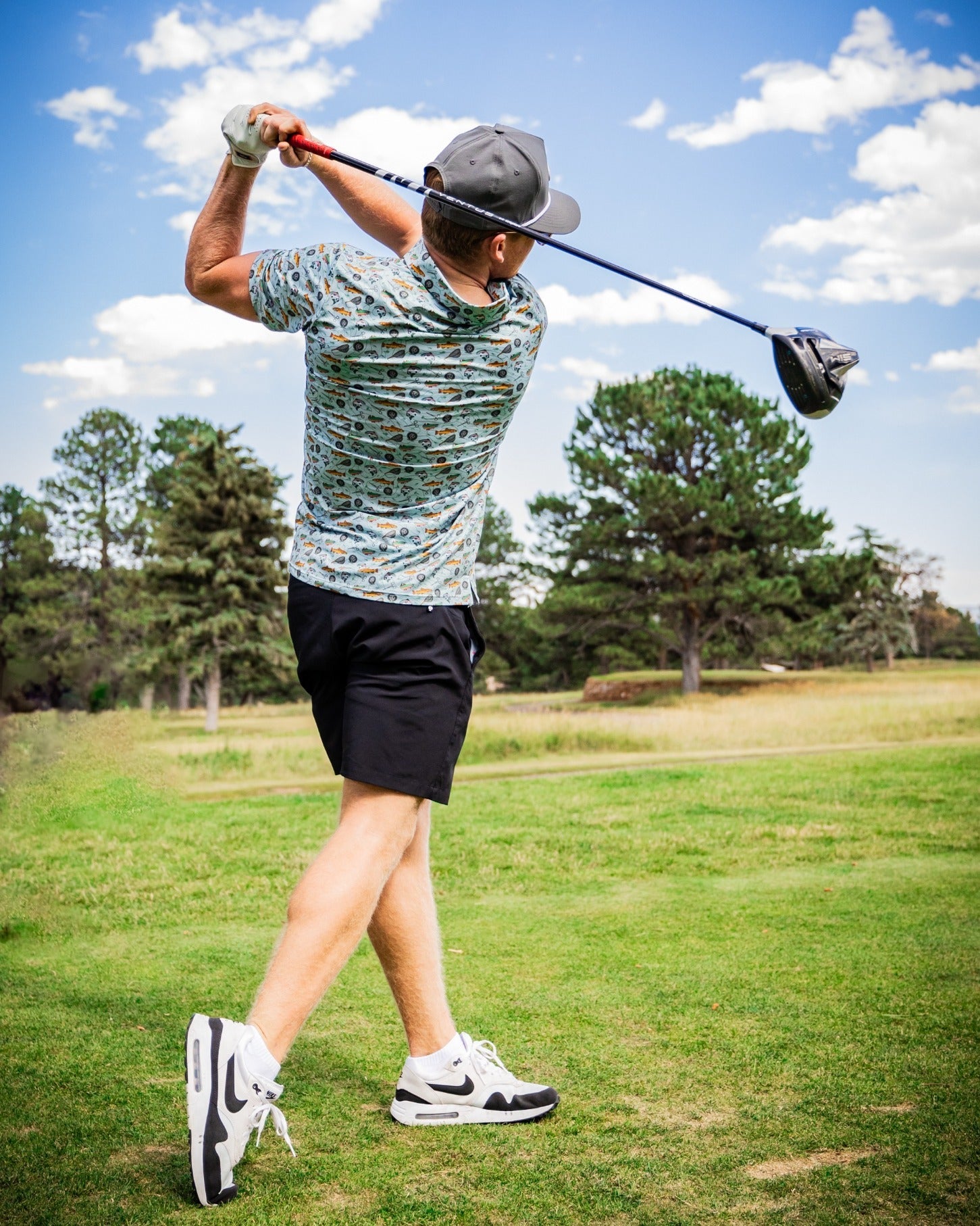 A golfer swings a driver on the course, wearing a colorful Catch and Release shirt and black shorts, showcasing bold style.