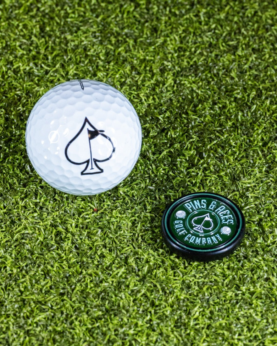 Green Pop Out Medallion Ball Marker next to a golf ball with an ace of spades design on a grassy surface.