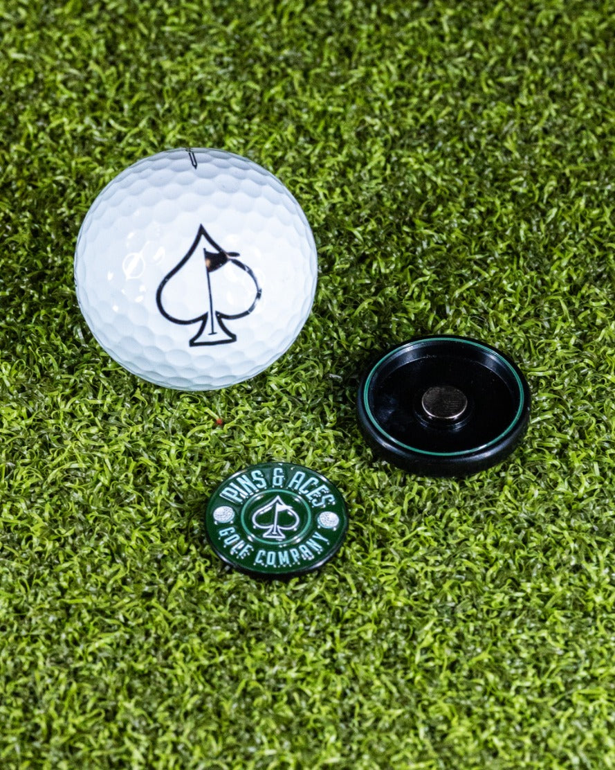 Green Pop Out Medallion Ball Marker next to a golf ball with an ace of spades logo, set on a grass background.