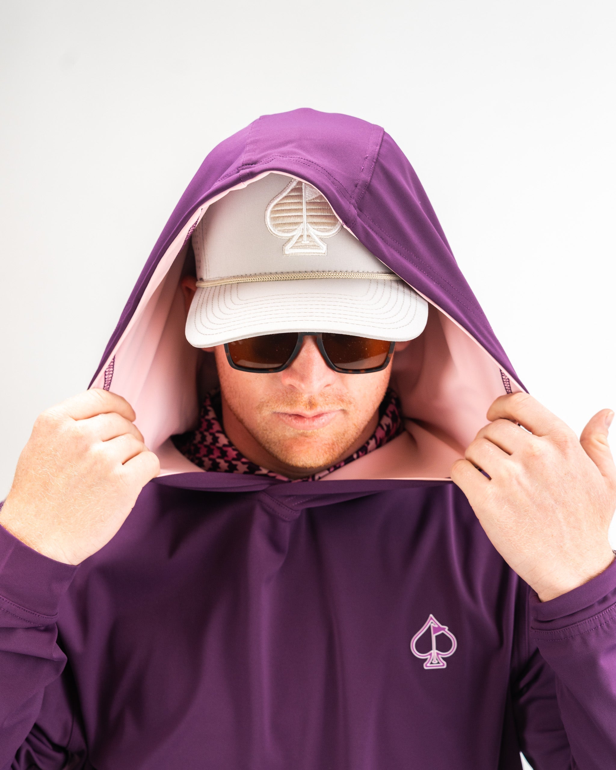 Man wearing a houndstooth raspberry performance golf hoodie with a hood up, sunglasses, and a cap, showcasing a stylish look.