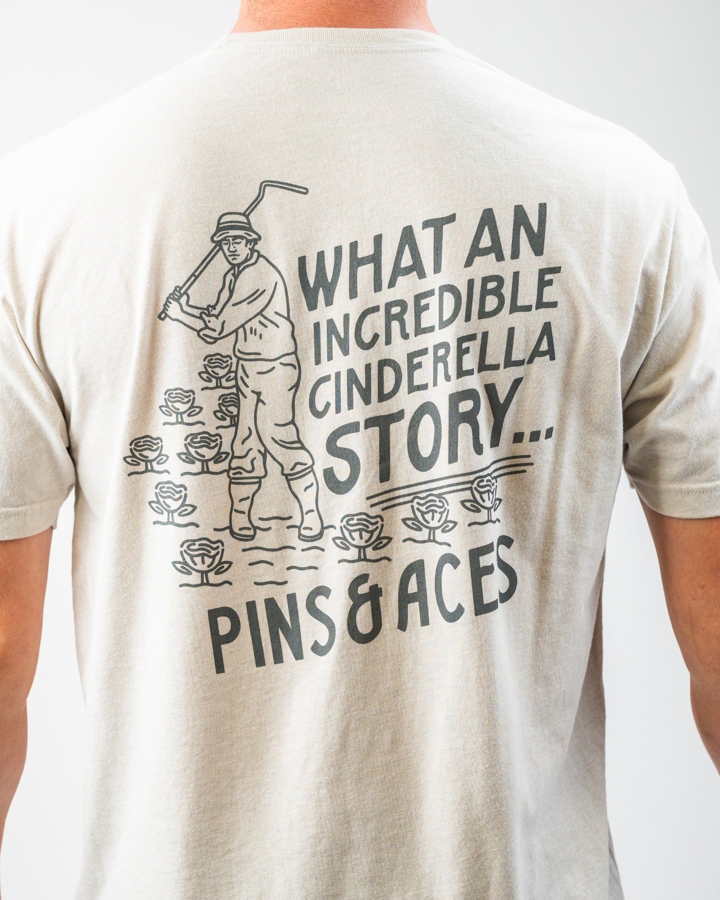 Back view of a light-colored tee shirt featuring a golfer and the text "What an incredible Cinderella story... Pins & Aces.