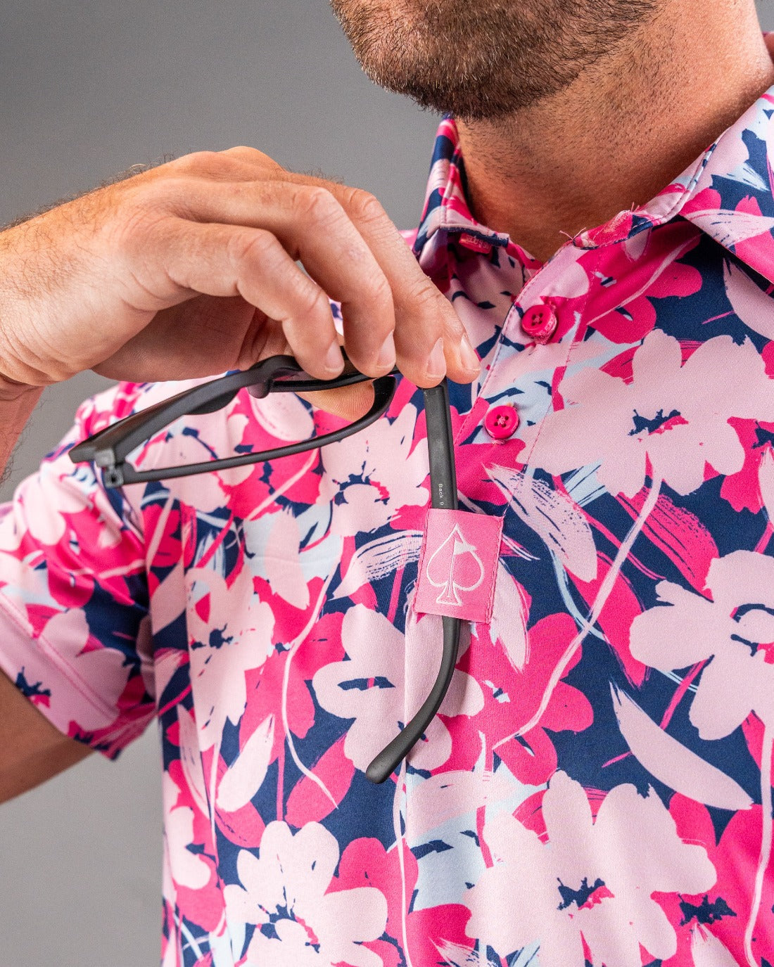 Close-up of a man in a vibrant pink, navy, and white floral golf shirt holding black sunglasses with a pink spade logo, sh...