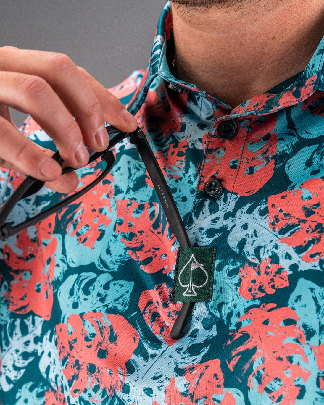Close-up of a golfer's colorful, floral-patterned shirt with a black spade logo tag, as they adjust stylish black sunglasses.