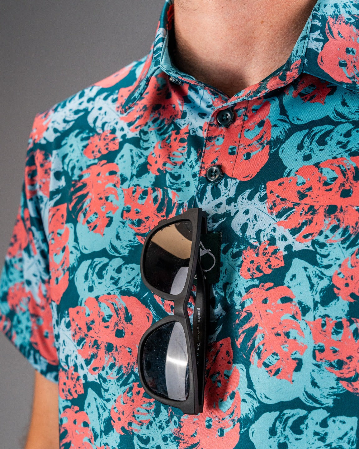 Bold floral-patterned golf shirt in blue, coral, and teal with sunglasses clipped to the chest for a standout golf look.