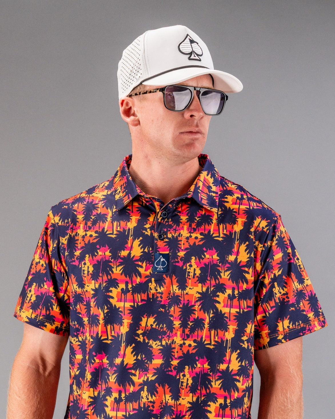 Man wearing bold, tropical palm tree patterned golf shirt, white cap with spade logo, and sunglasses, showcasing standout ...