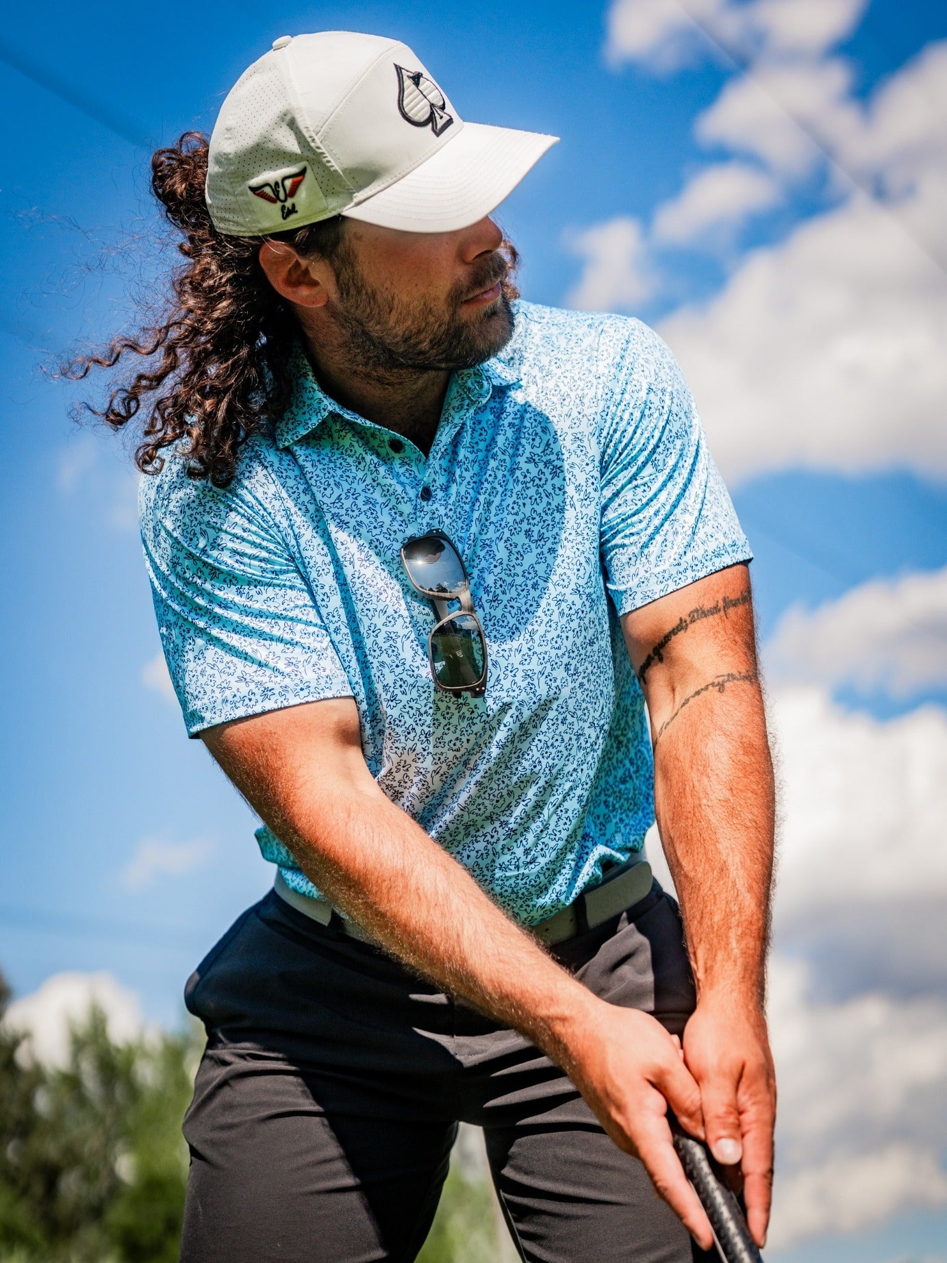 A golfer in a blue floral-patterned polo shirt and cap prepares to swing on a sunny day, showcasing bold golf style.