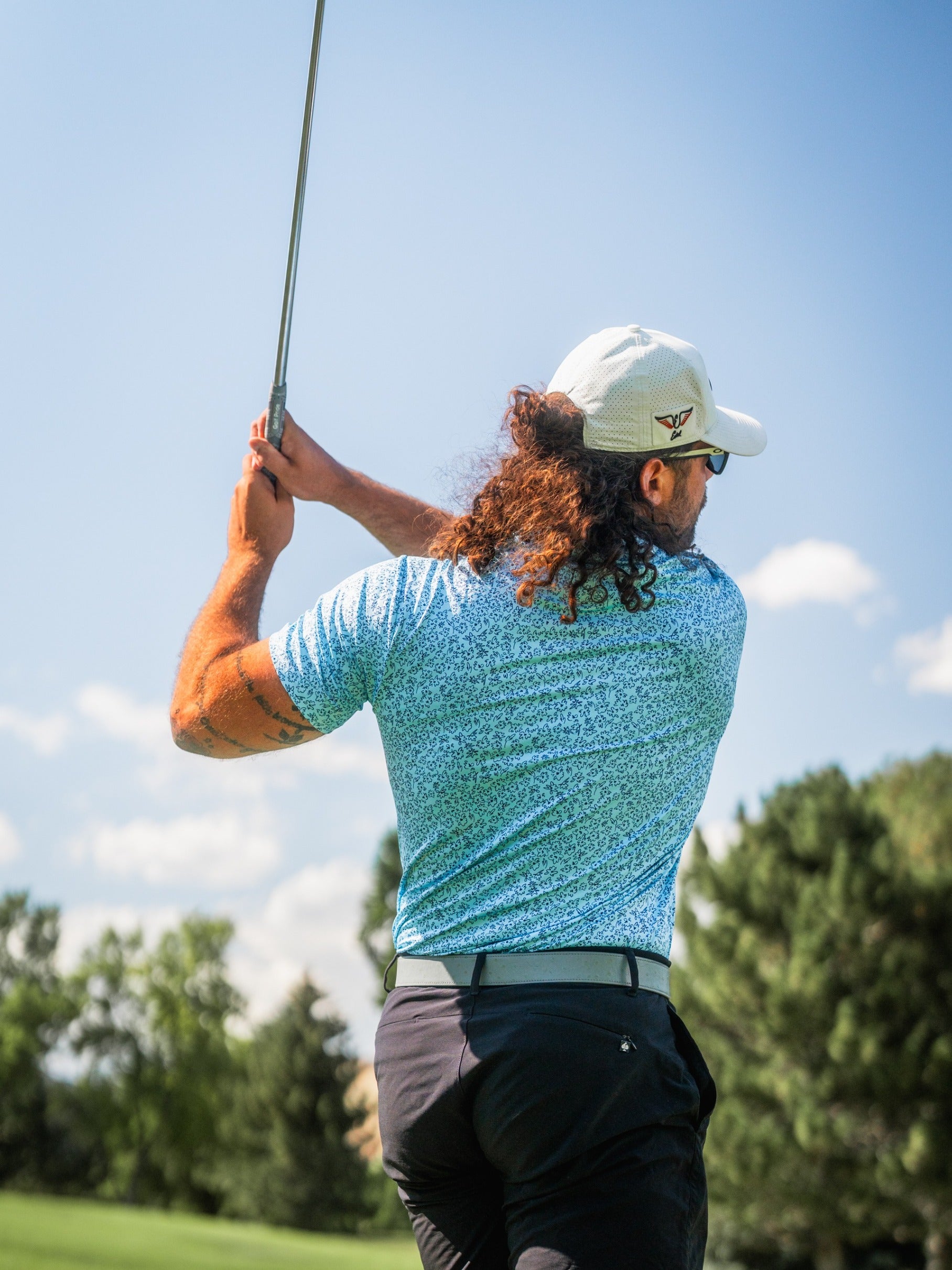 Man swinging a golf club, wearing a blue floral-patterned shirt and a cap, against a sunny golf course backdrop.