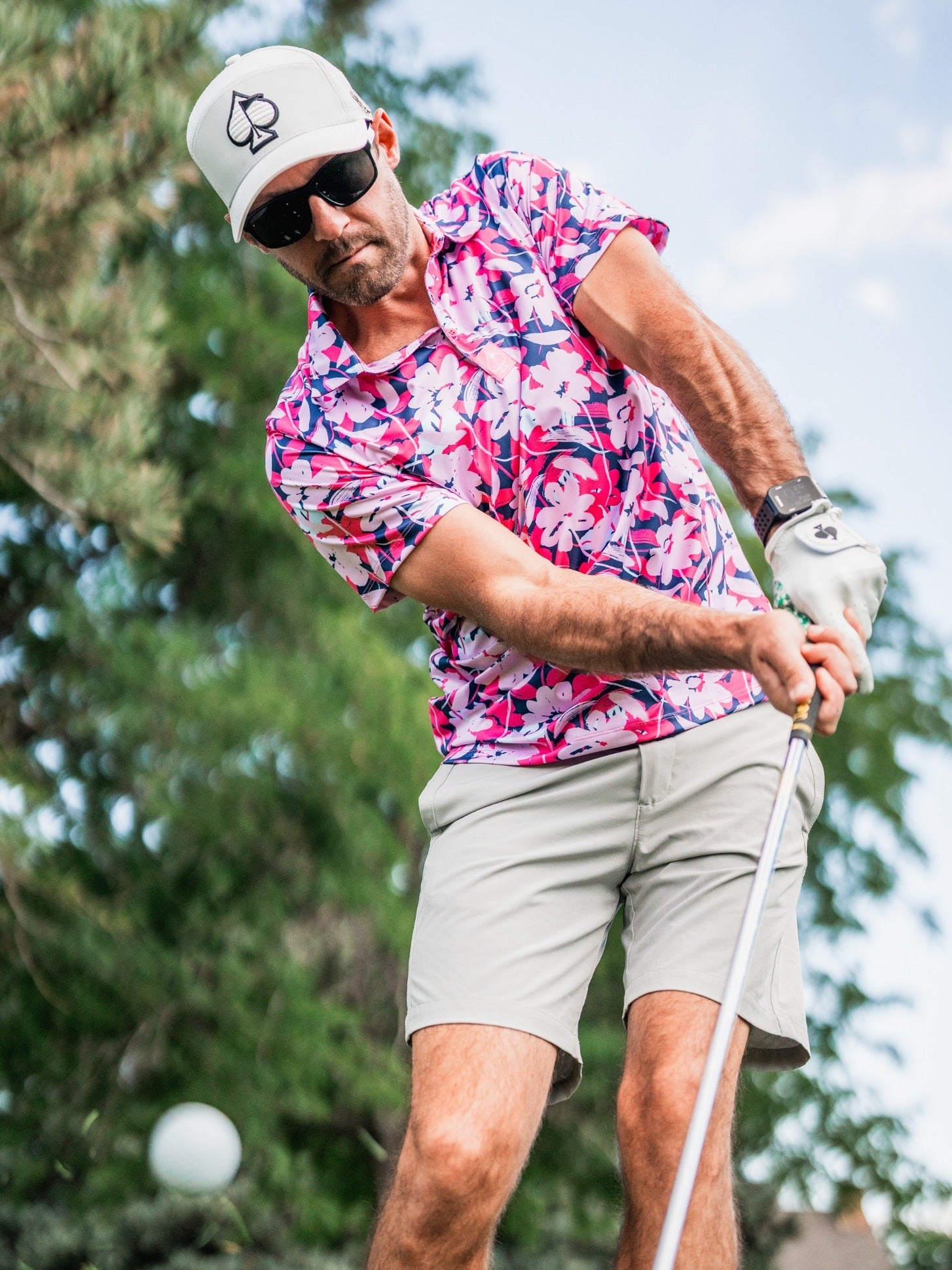 Man in a pink floral golf shirt, white cap with spade logo, black sunglasses, and white glove, preparing to swing golf clu...
