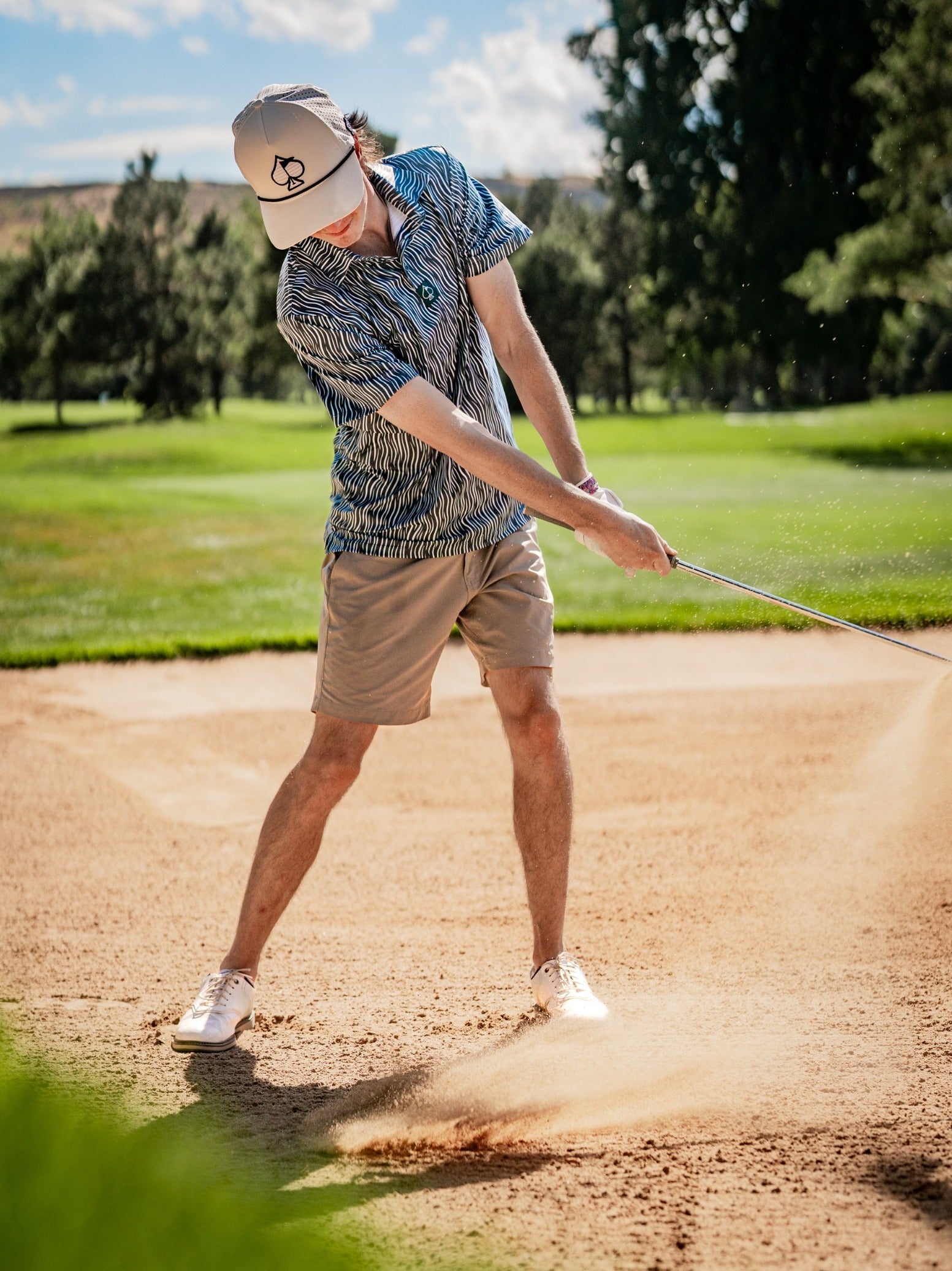 Golf player in a striped shirt, beige shorts, and white cap hitting a shot from a sand bunker on a sunny course.