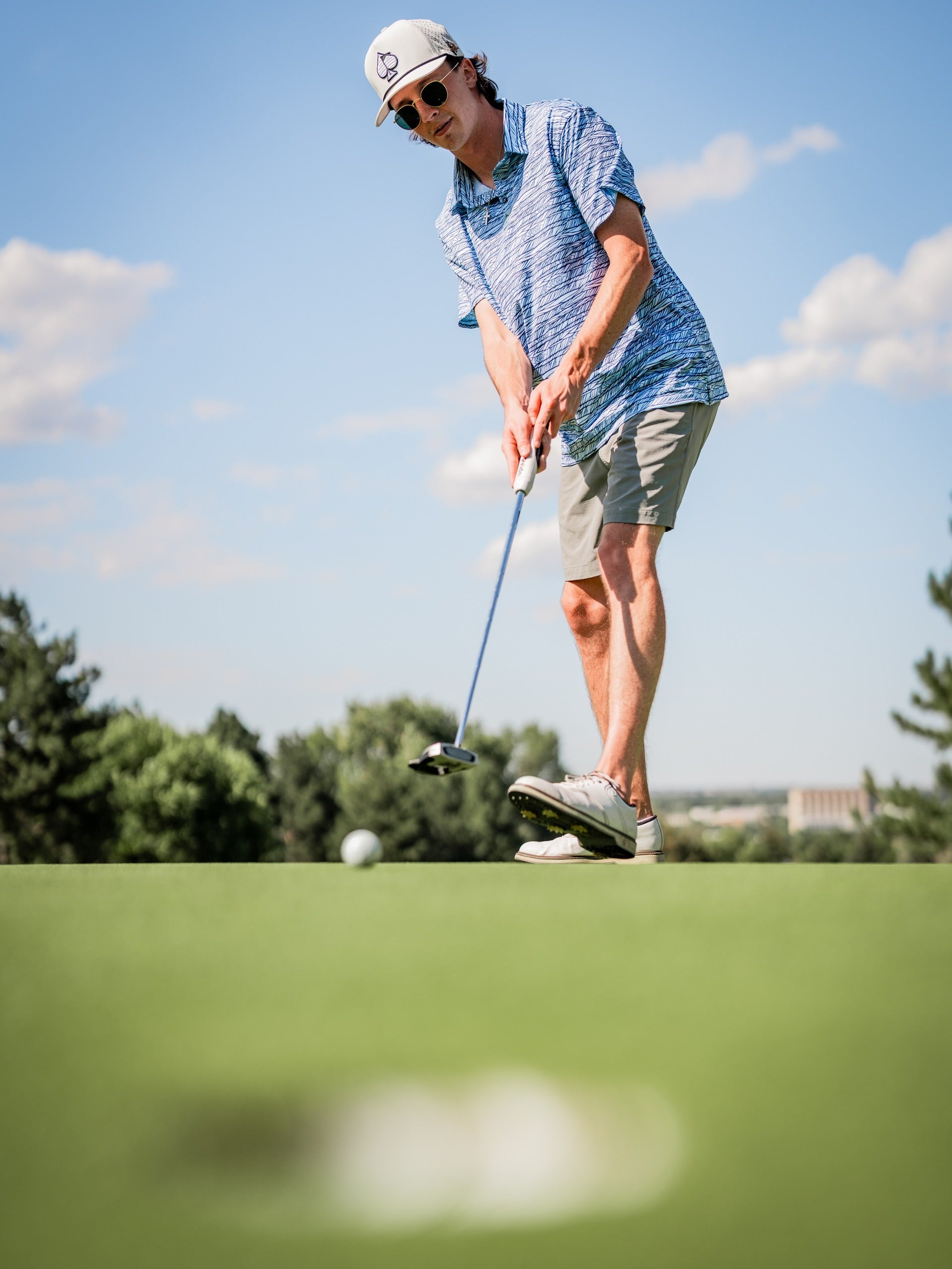 Male golfer in a blue patterned shirt, beige shorts, white cap, and sunglasses, putting on a sunny golf course.