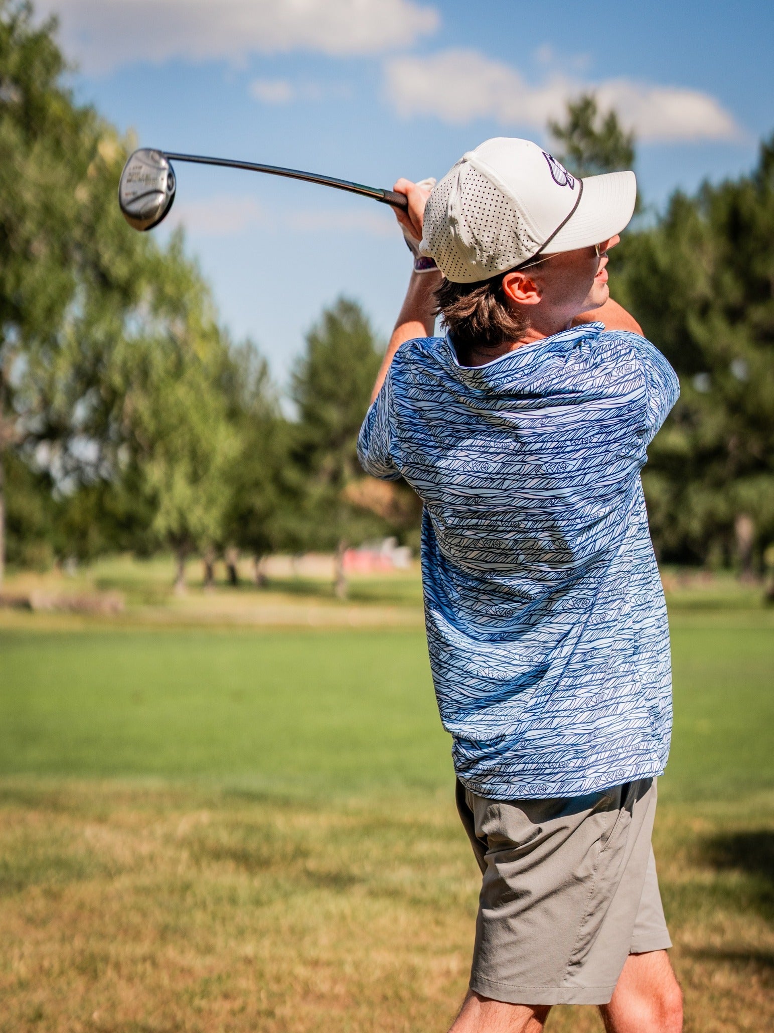 Young man in a blue patterned golf shirt and beige shorts swings a golf club on a sunny course with trees and blue sky in ...