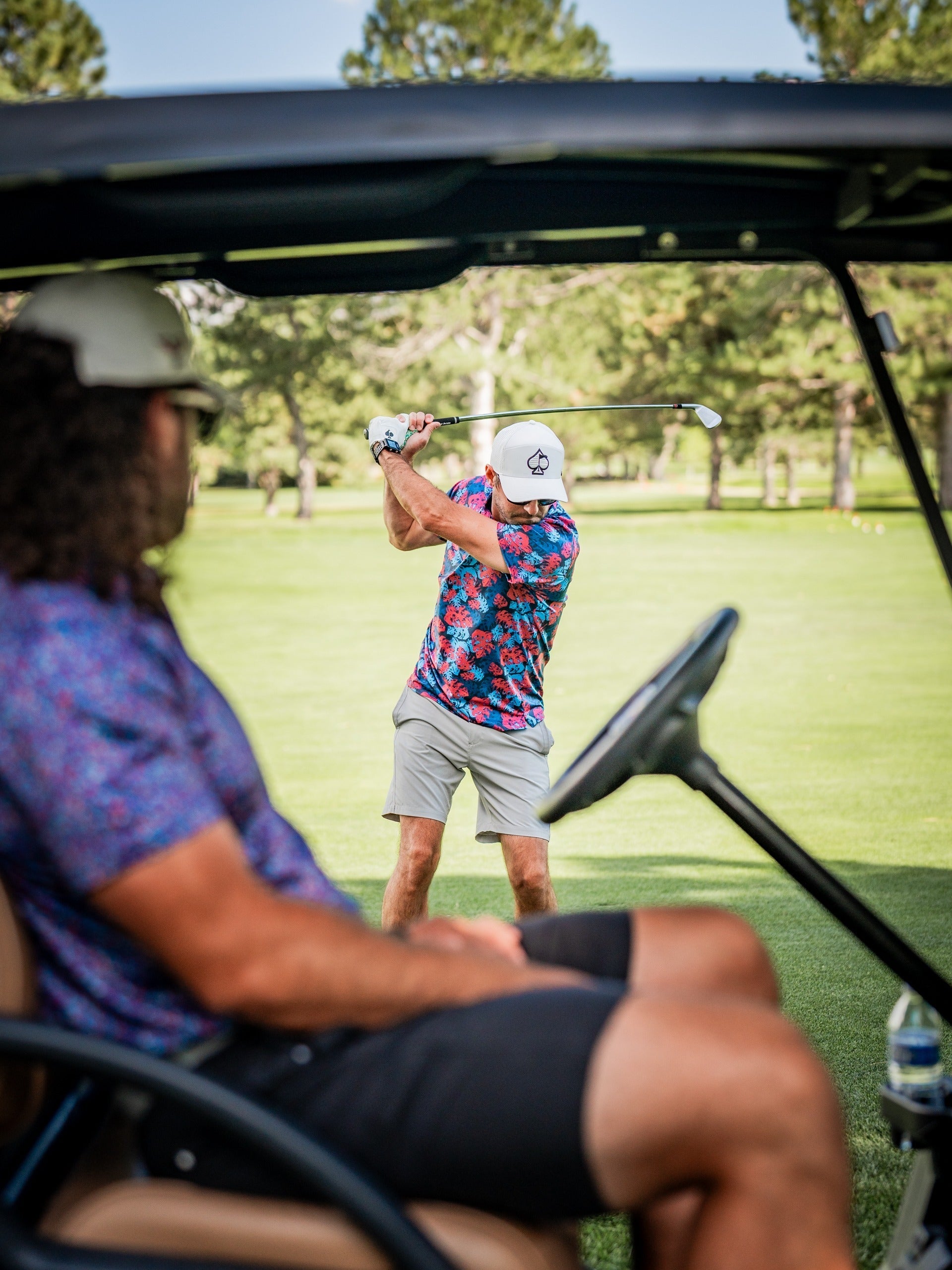 Two golfers in colorful, bold golf apparel on a lush green course, one swinging a club, the other seated in a golf cart.