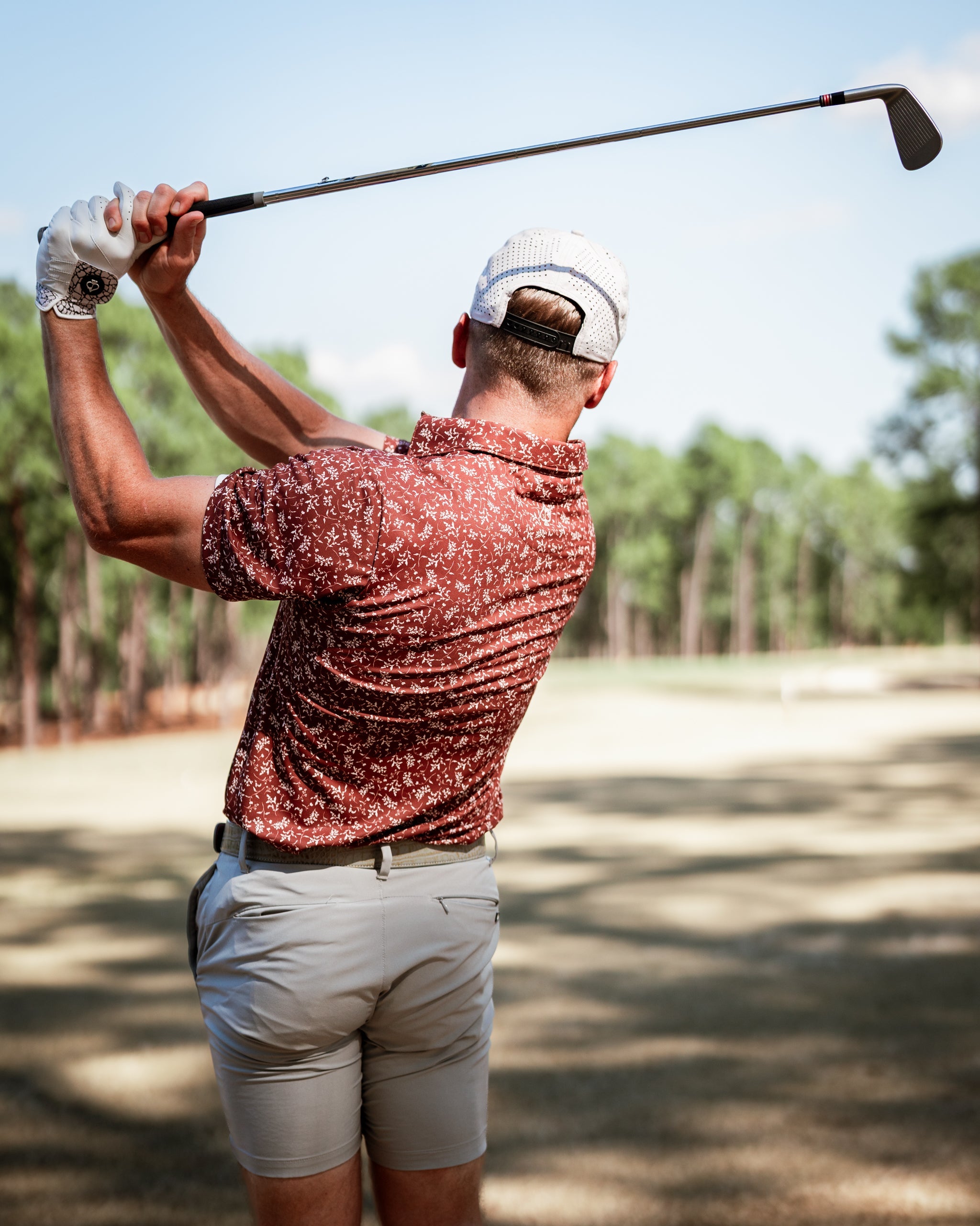 A golfer swings a club wearing a floral-patterned shirt and light shorts on a sunny course surrounded by trees.