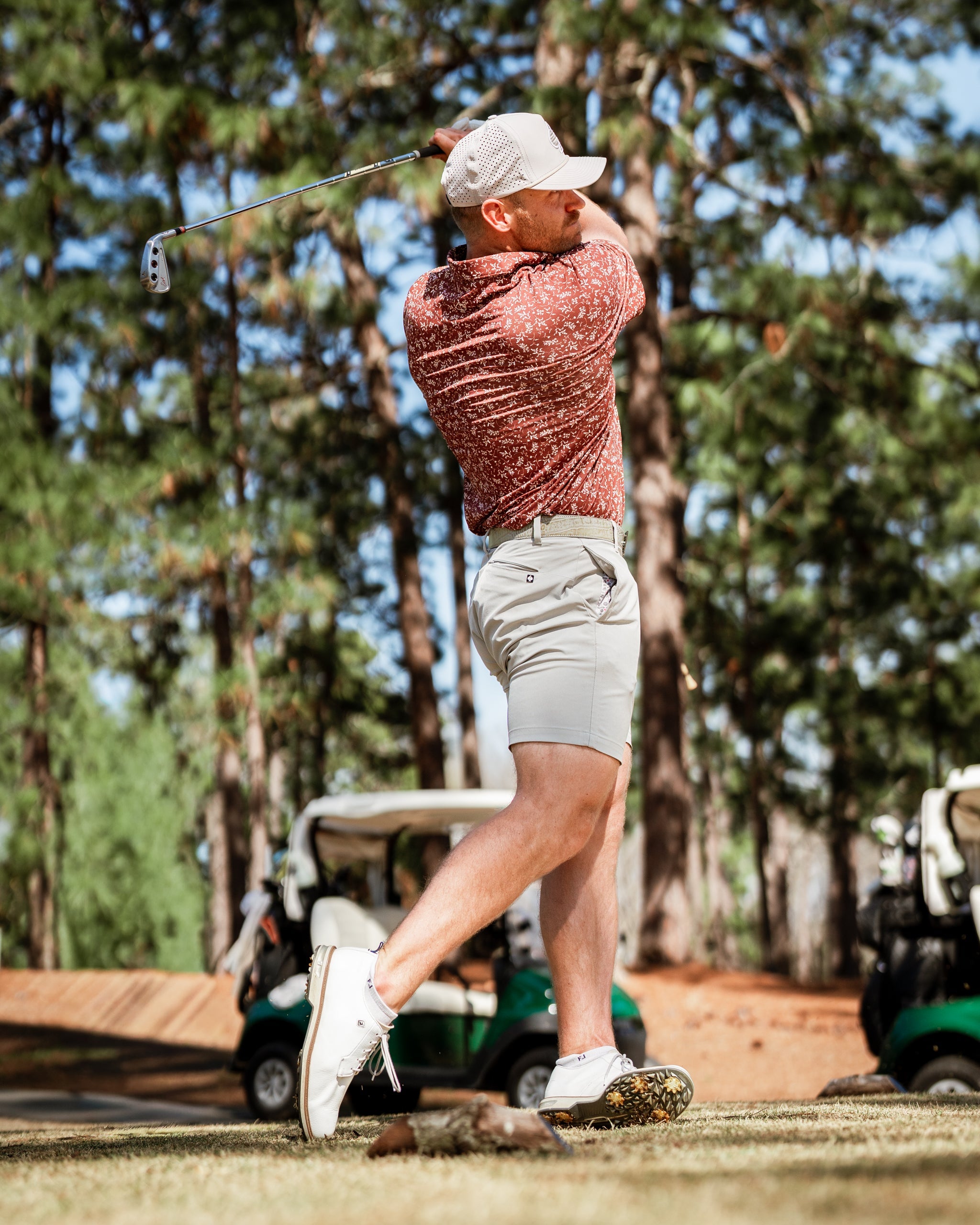 A golfer swings a club in a floral-patterned shirt and shorts, showcasing vibrant autumn colors on the course.