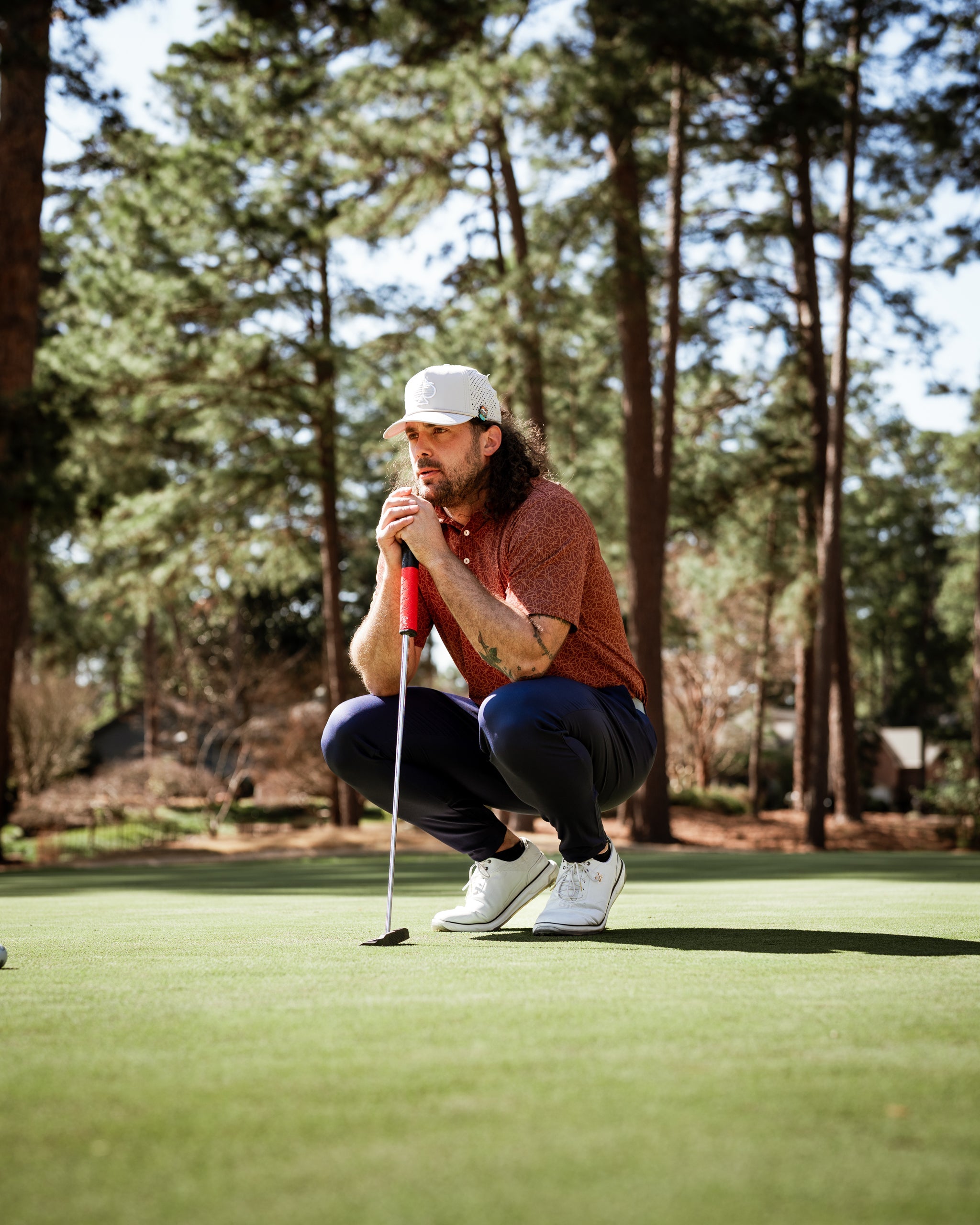 A golfer in a stylish orange shirt and white cap crouches on the green, focusing intently on his putt.