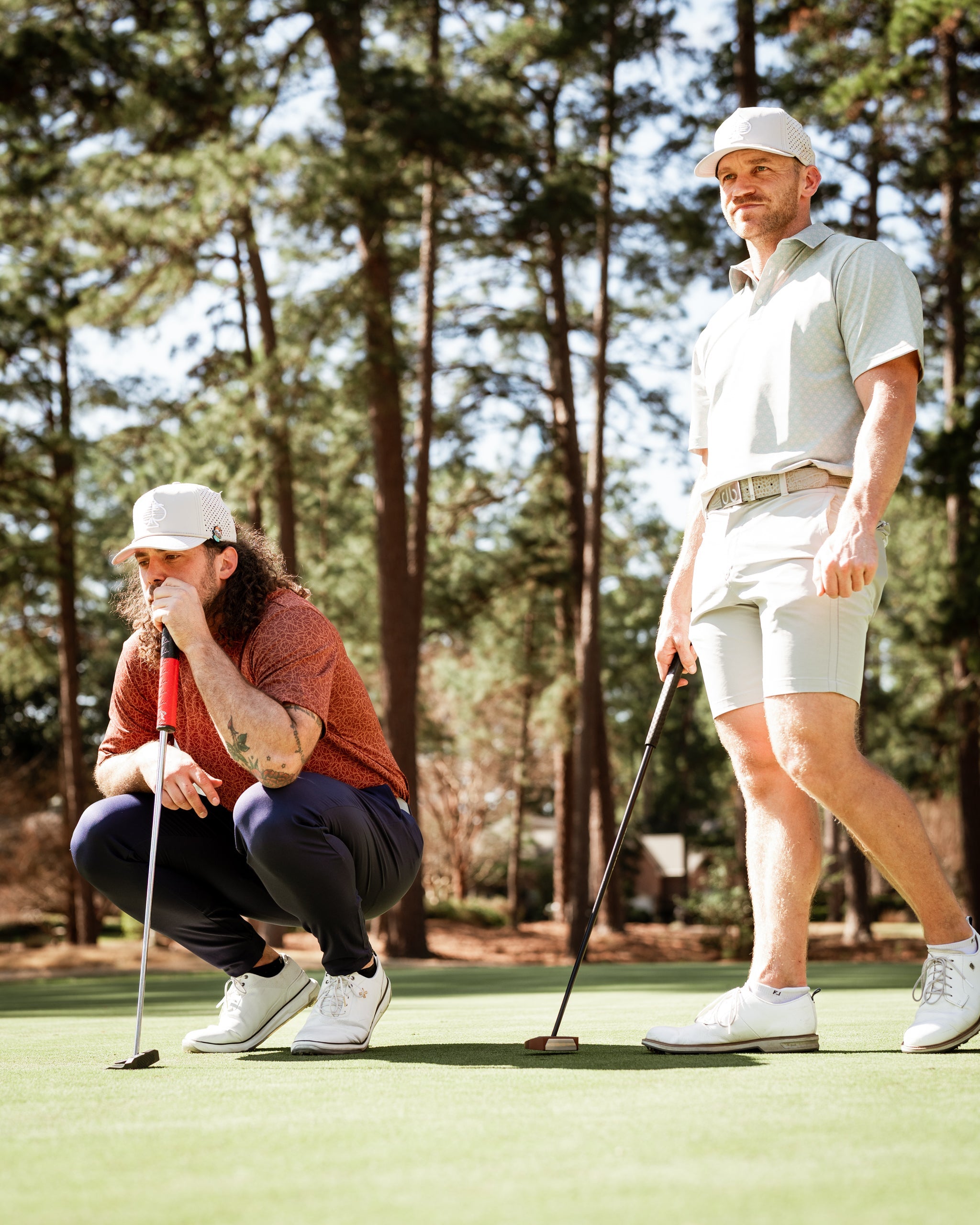 Two golfers on a putting green, one crouching to line up a shot while the other stands nearby, both in stylish golf apparel.
