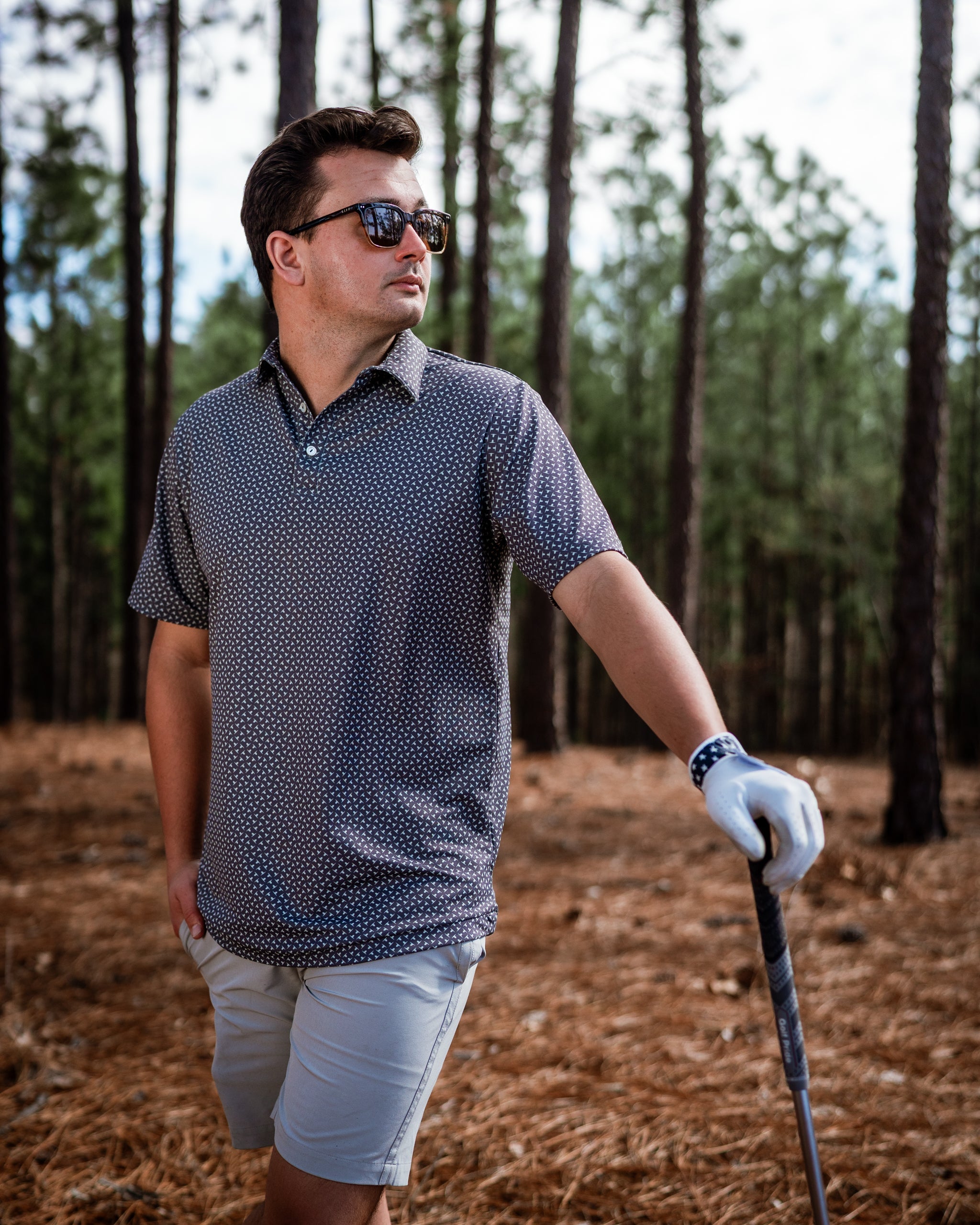 Stylish golfer wearing a patterned polo shirt and shorts, posing confidently with a golf club in a wooded setting.