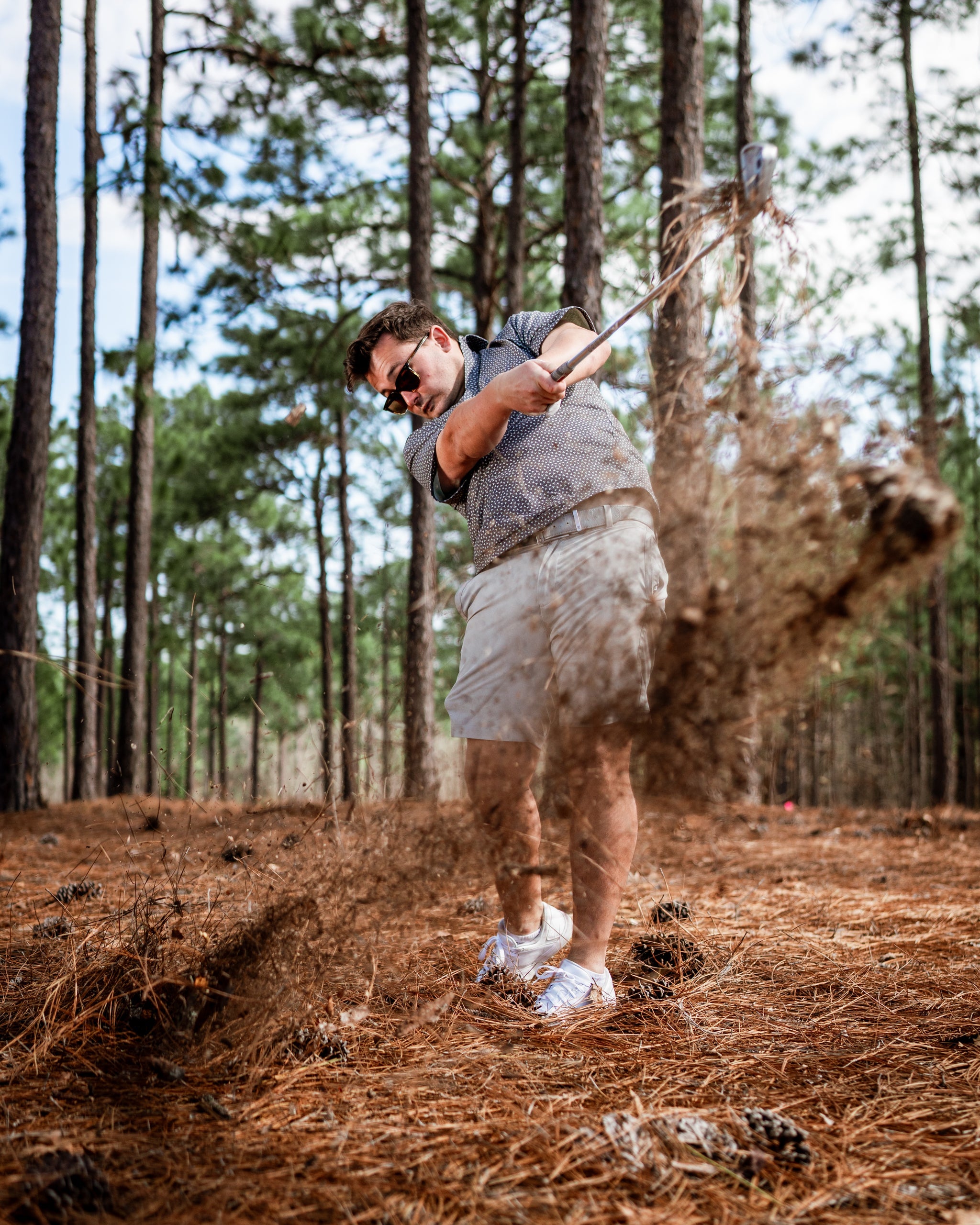 A golfer swings a club in a forested area, sending dirt and pine needles flying as he makes contact with the ground.