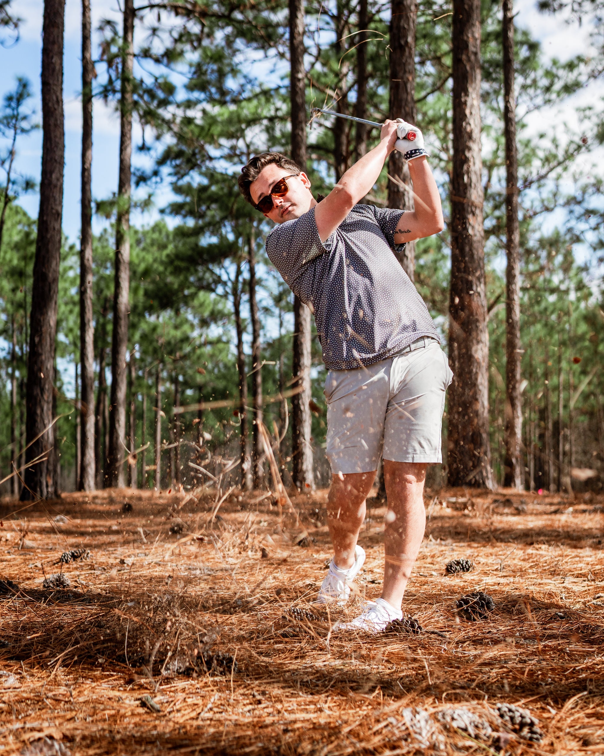 A golfer swings a club in a forested area, showcasing the Jade Drops golf apparel with a stylish and relaxed look.