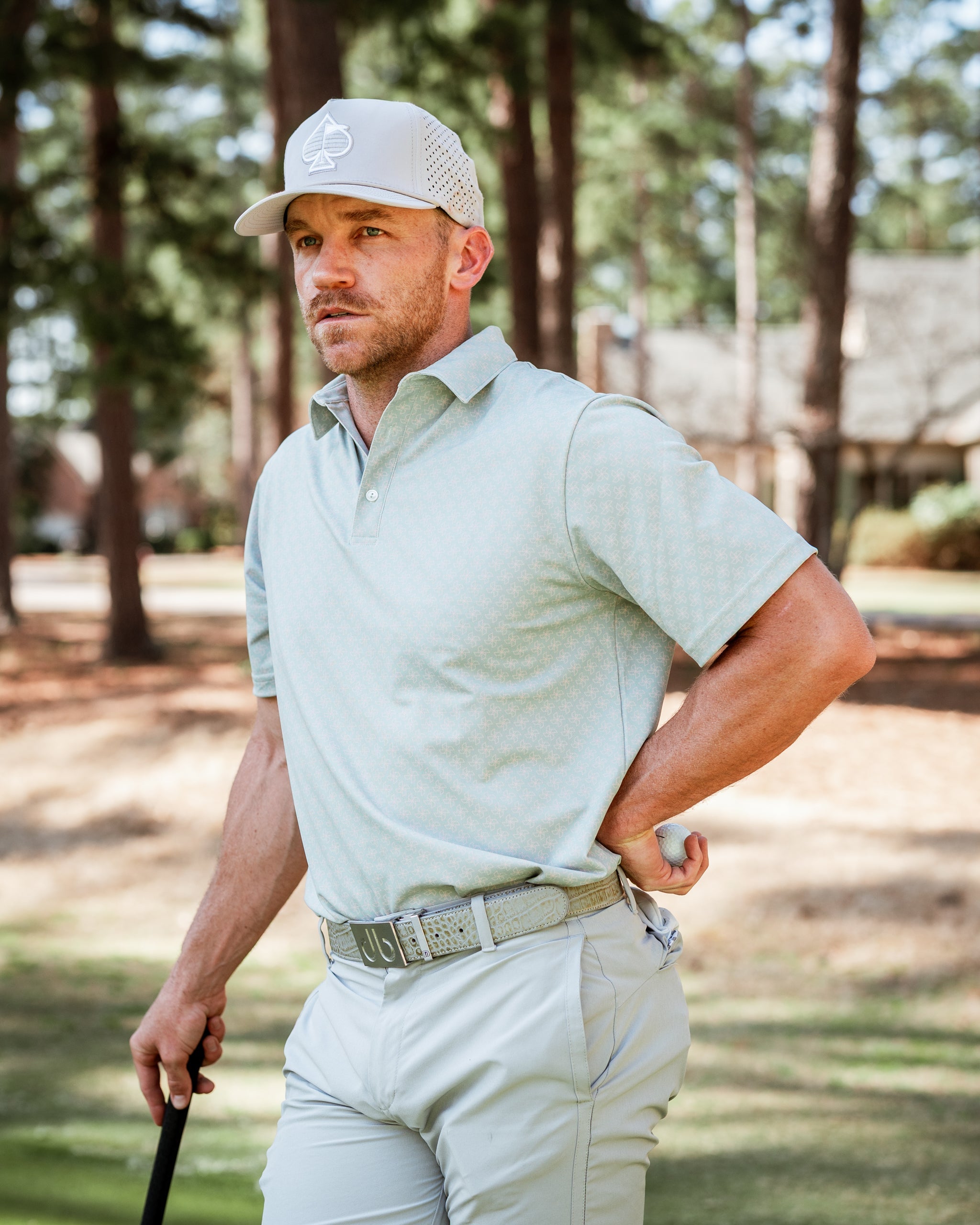 Man wearing a light green origami-patterned golf polo and a gray cap, standing on a golf course with trees in the background.